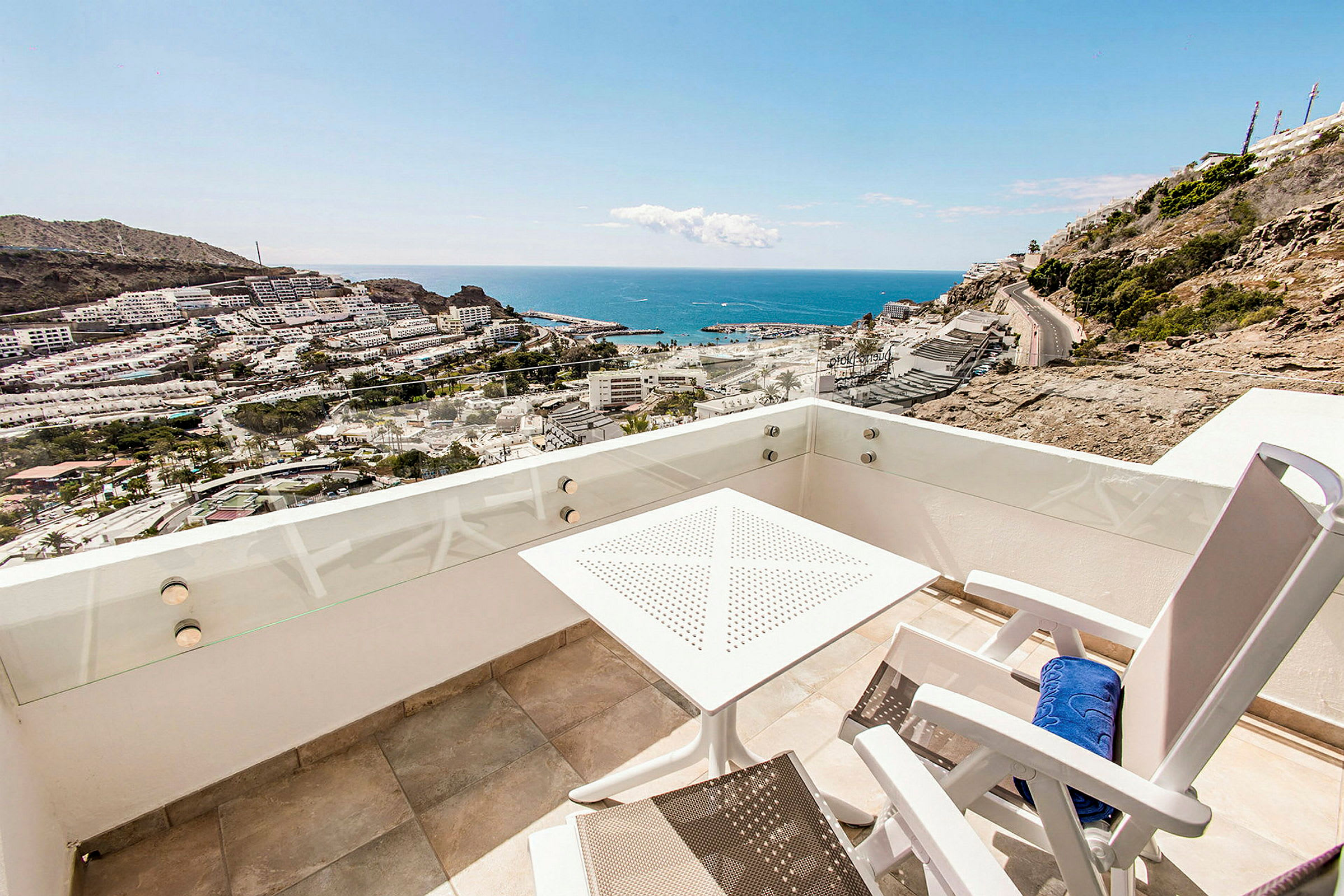 Udsigt fra en balkon med et bord og stole mod havet og en by. (AI-genereret tekst)