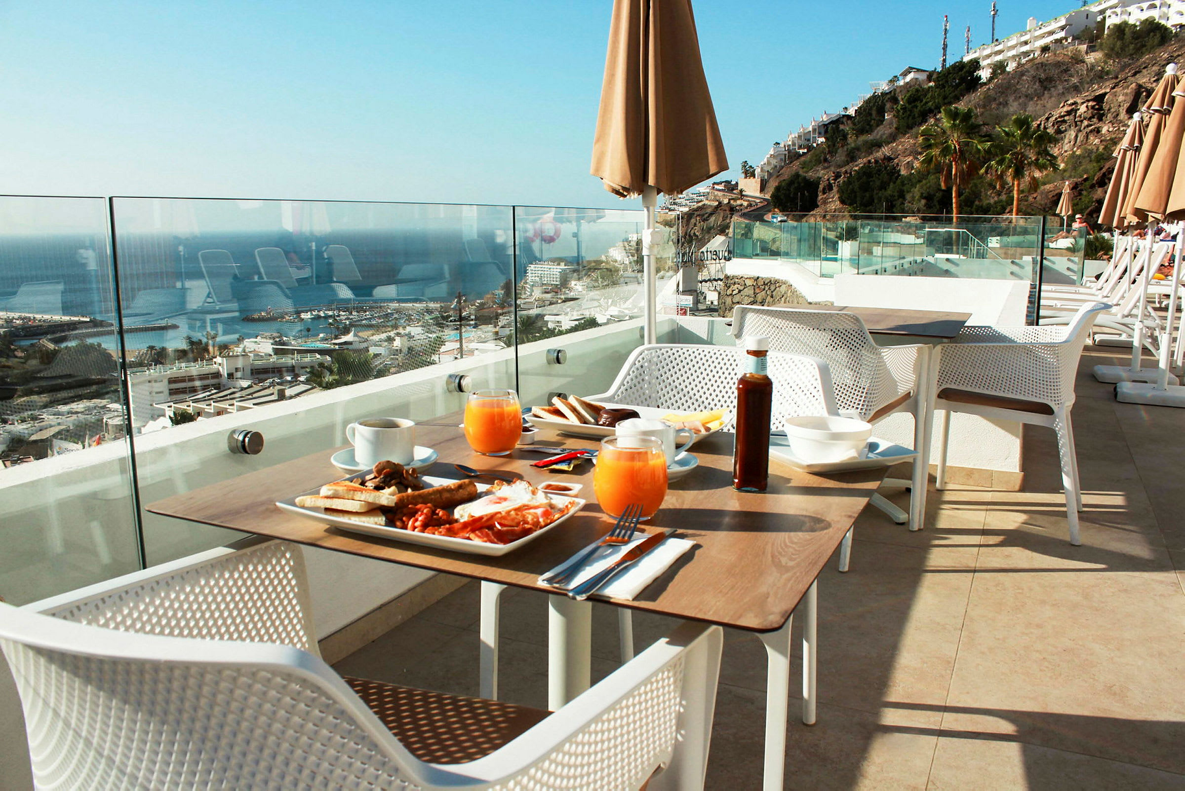Udsigt over et morgenmadsbord på en terrasse med glasrækværk, mod havet og en klippe med bygninger. (AI-genereret tekst)