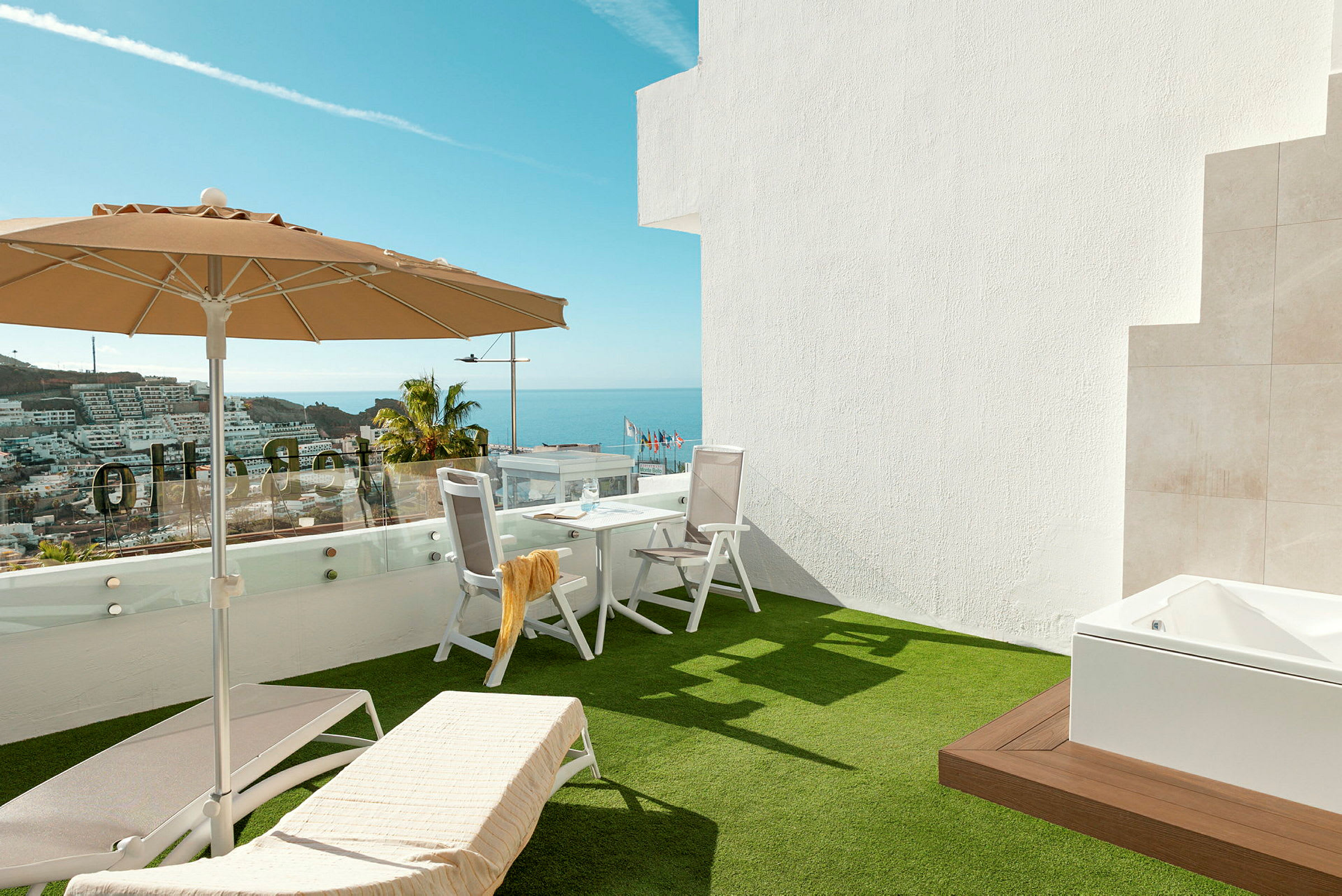 Terrasse med solsenge, parasol og bord med stole, havudsigt i baggrunden. (AI-genereret tekst)