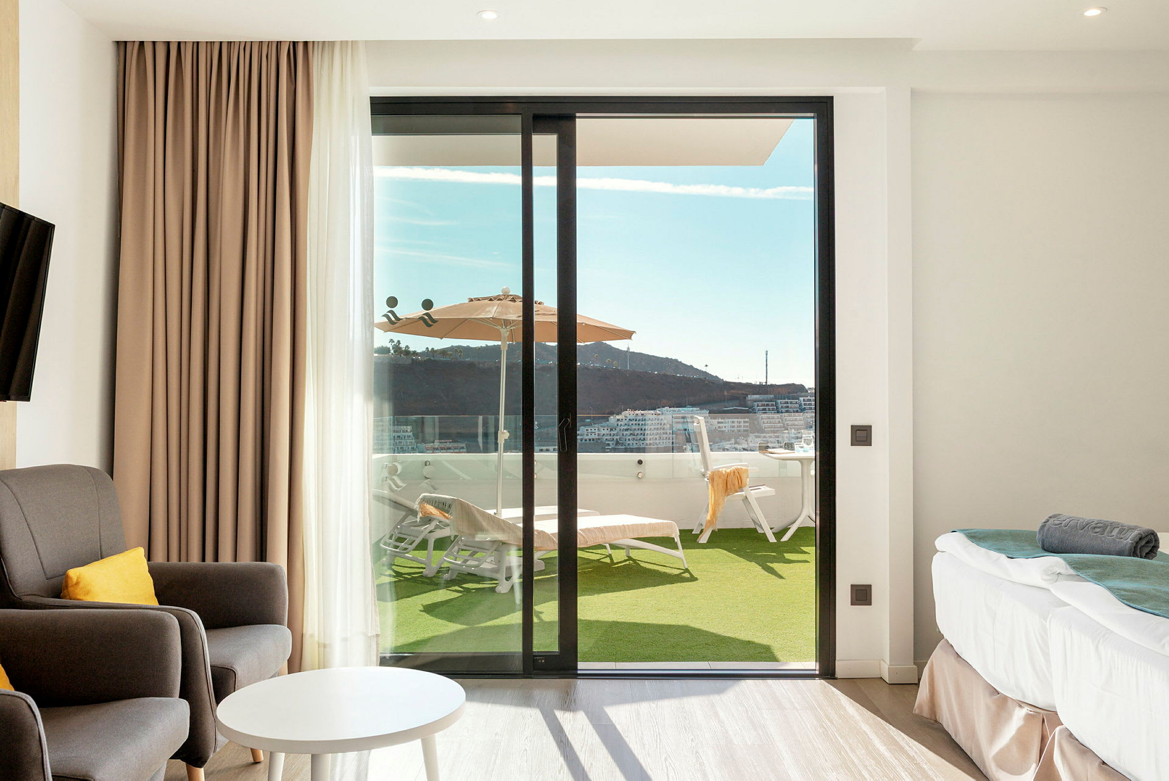Et hotelværelse med siddepladser, bord og seng, samt en glasdør der fører ud til en terrasse med liggestole og parasol. (AI-genereret tekst)