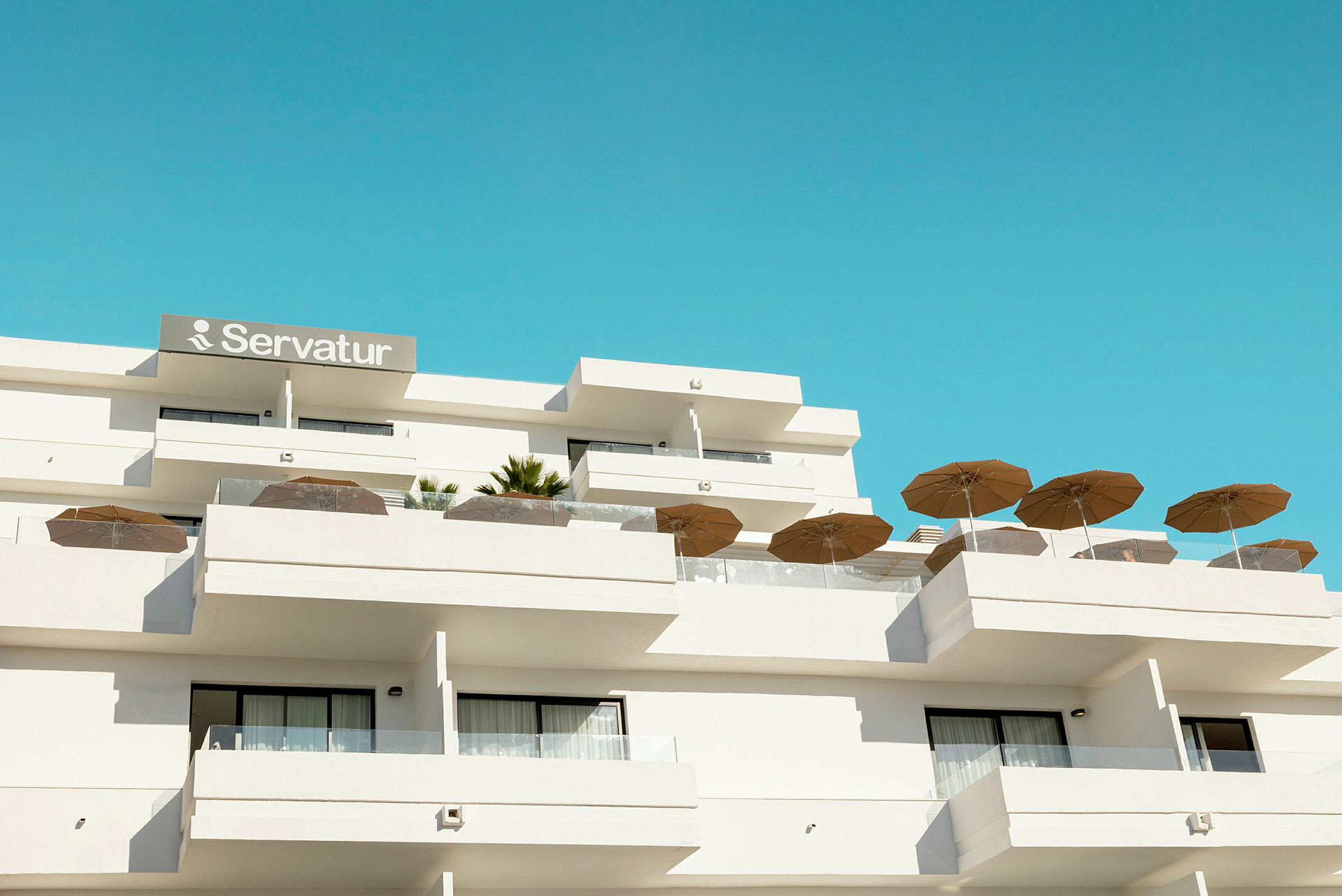 Hotelbygning med balkoner og parasoller mod en blå himmel. (AI-genereret tekst)