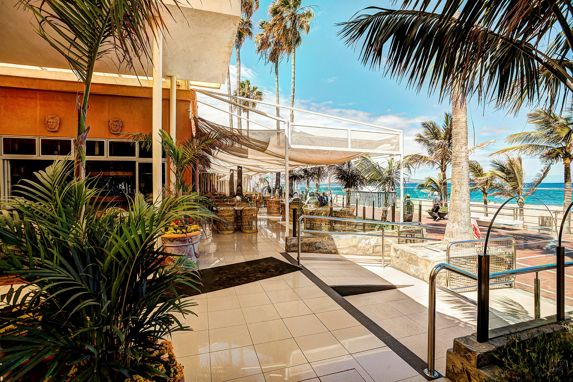 Restaurant ved strandpromenade med palmer og hav i baggrunden. (AI-genereret tekst)