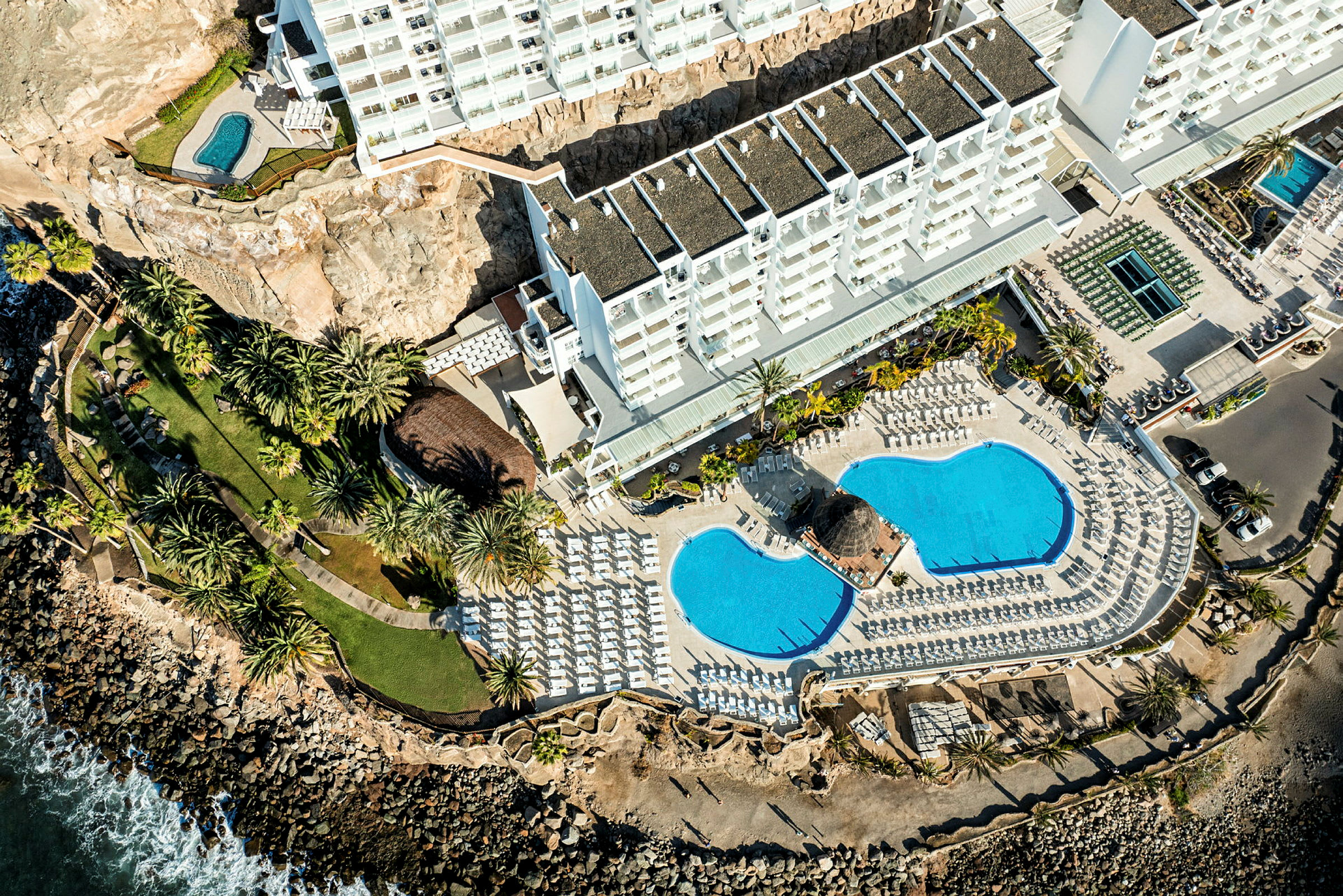 Luftfoto af hotelområde med bygninger, pools, grønne områder og kystlinie. (AI-genereret tekst)