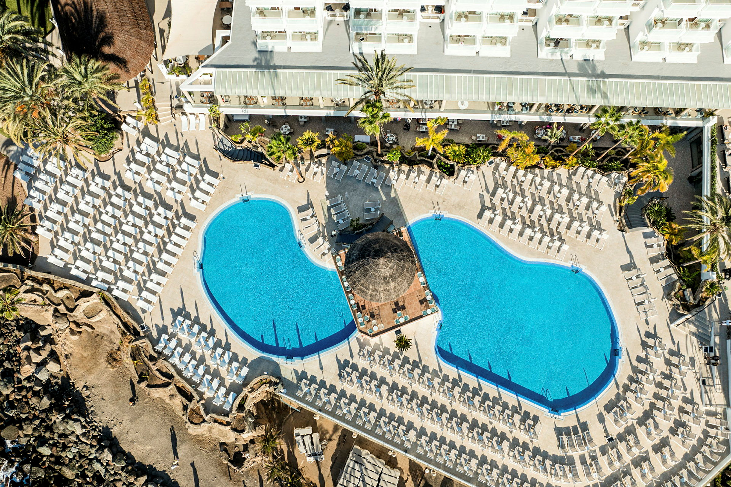 Fugleperspektiv af et hotelpoolområde med to pools og mange liggestole. (AI-genereret tekst)
