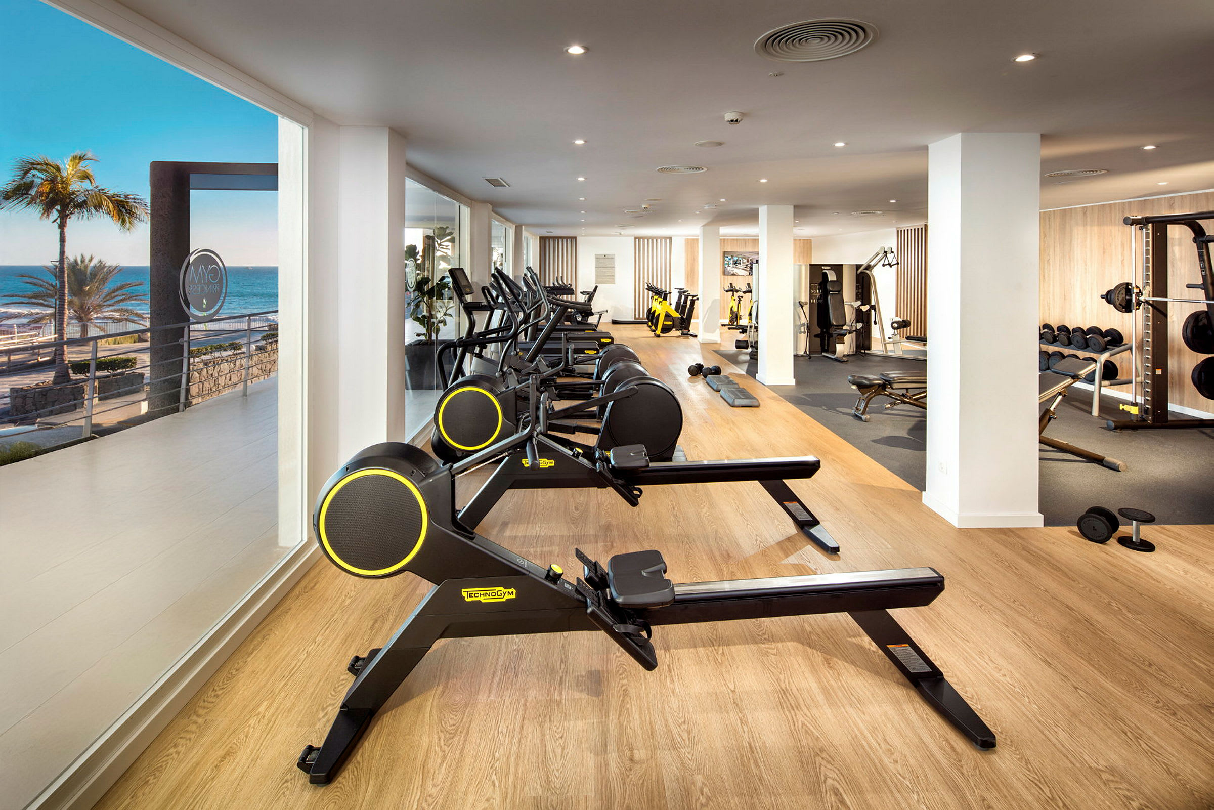 Fitnesscenter med træningsmaskiner og havudsigt gennem vinduet. (AI-genereret tekst)