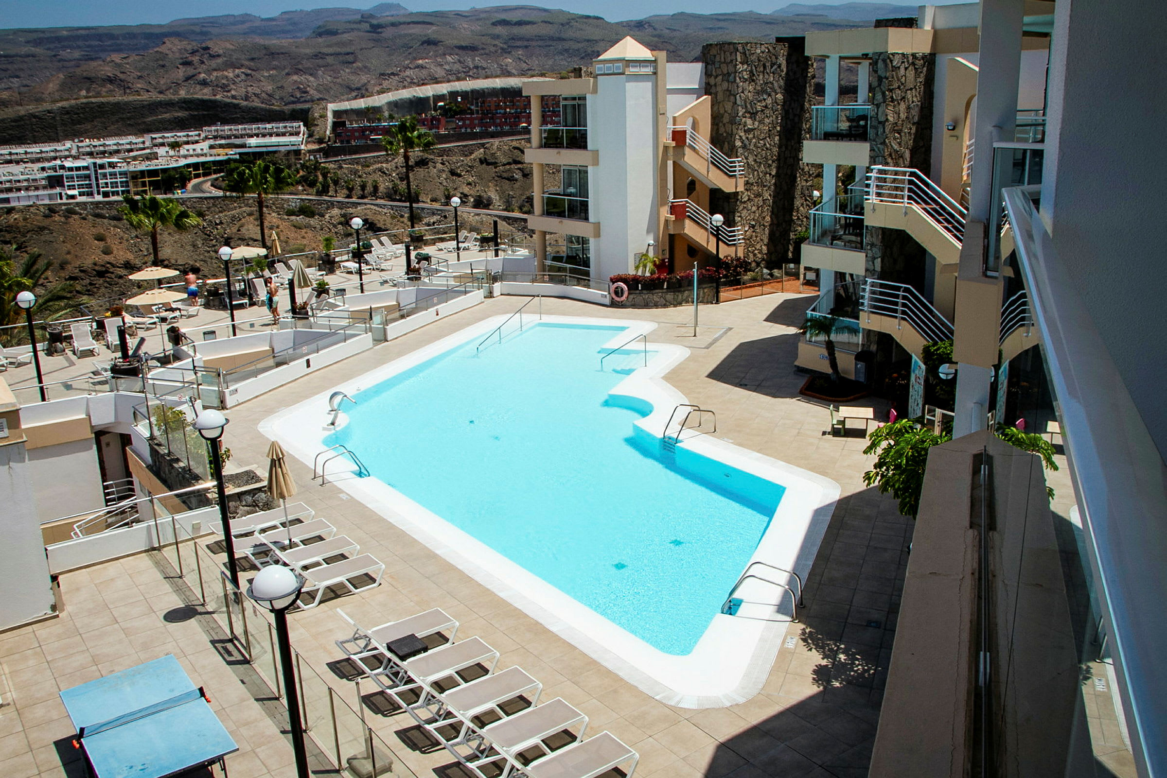 Billede af et hotelområde med udendørs swimmingpool, liggestole og omkringliggende bygninger. (AI-genereret tekst)
