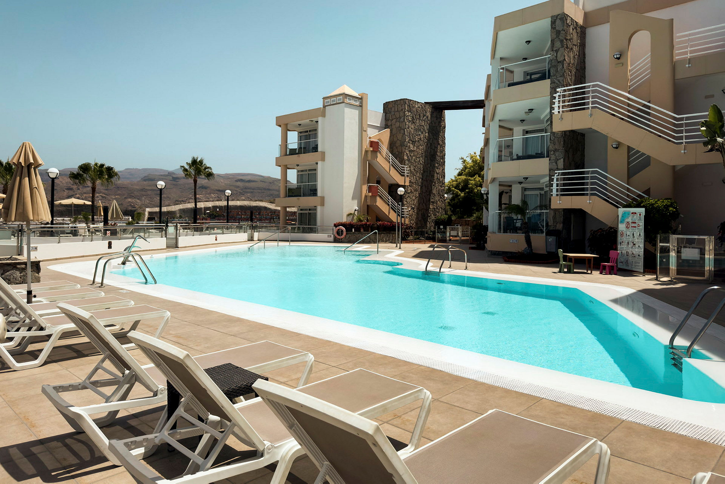 Udendørs pool ved hotel med liggestole og bygninger i baggrunden. (AI-genereret tekst)