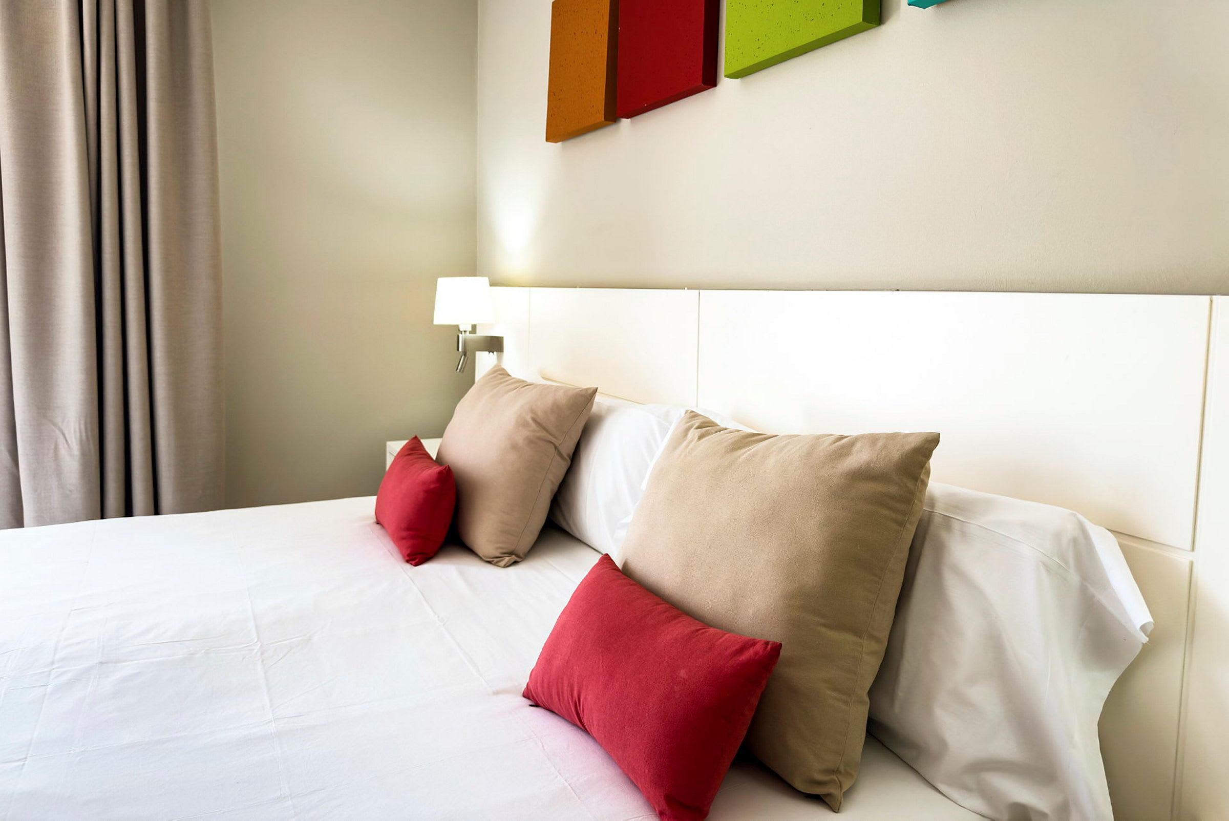 Hotelværelse med en seng redt med hvide lagner og flere puder i røde og beige farver. (AI-genereret tekst)
