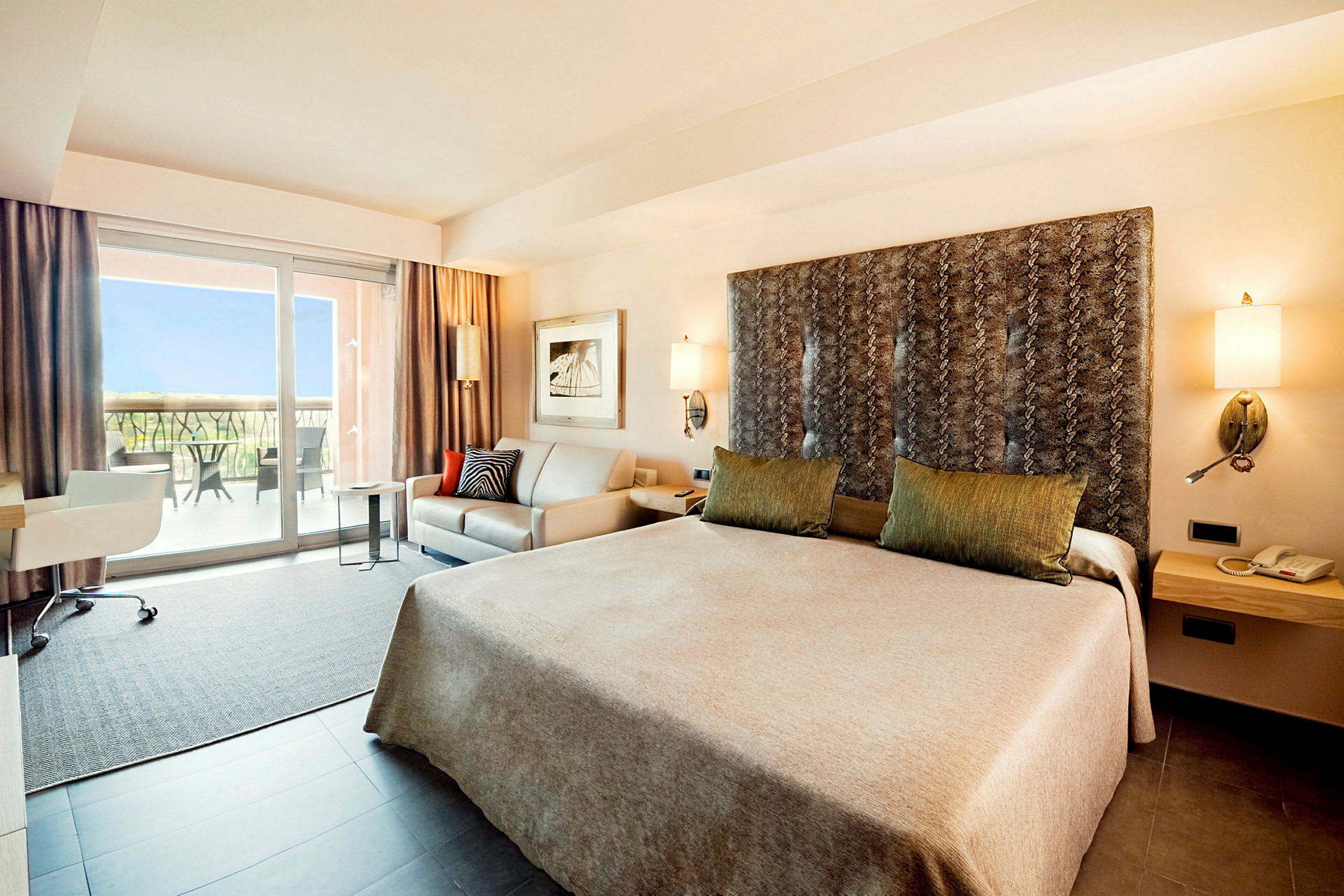 Et hotelværelse med en stor seng, sofa, skrivebord og et vindue, der fører til en balkon. (AI-genereret tekst)