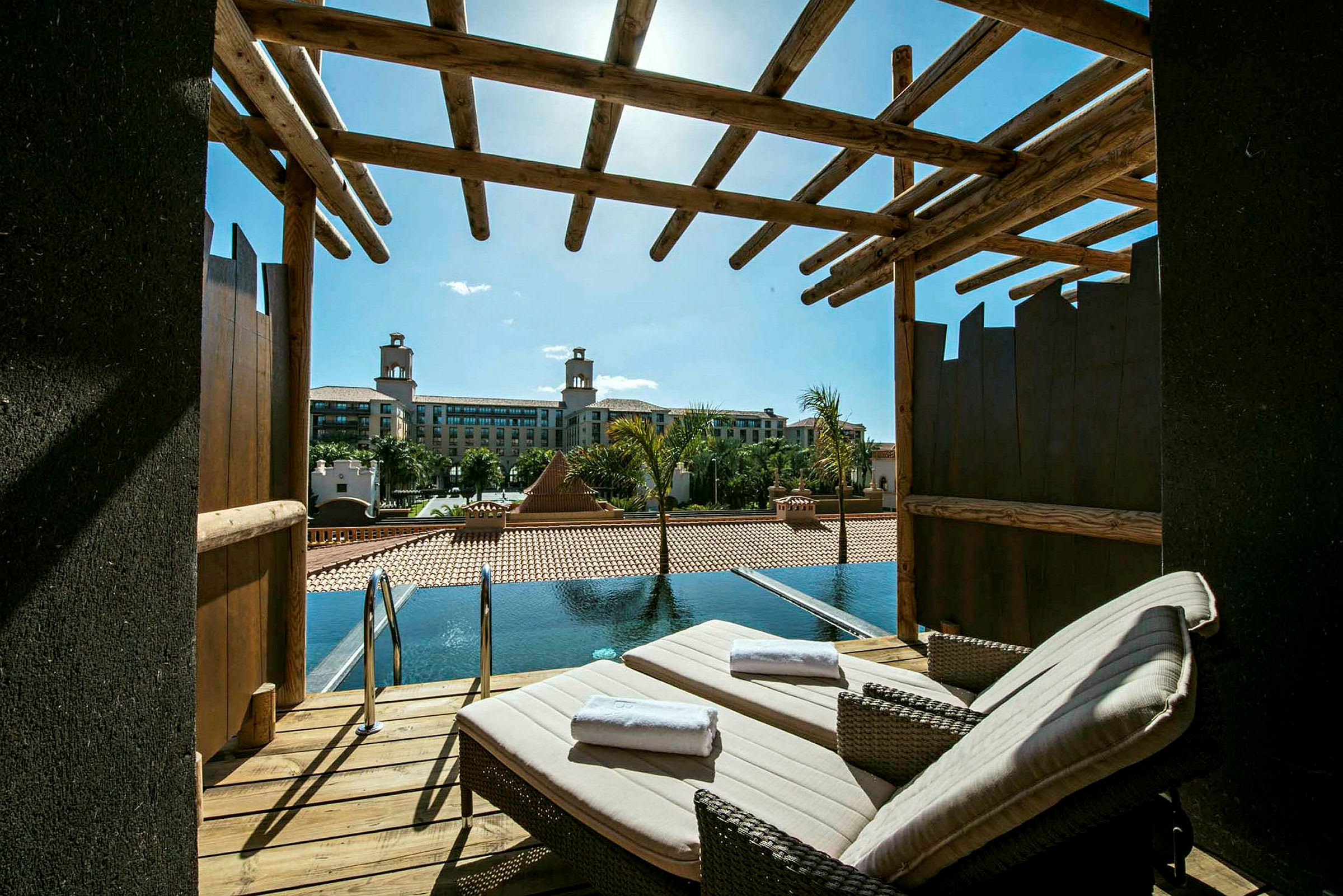 En terrasse med to liggestole og udsigt til en pool og et hotel i baggrunden. (AI-genereret tekst)