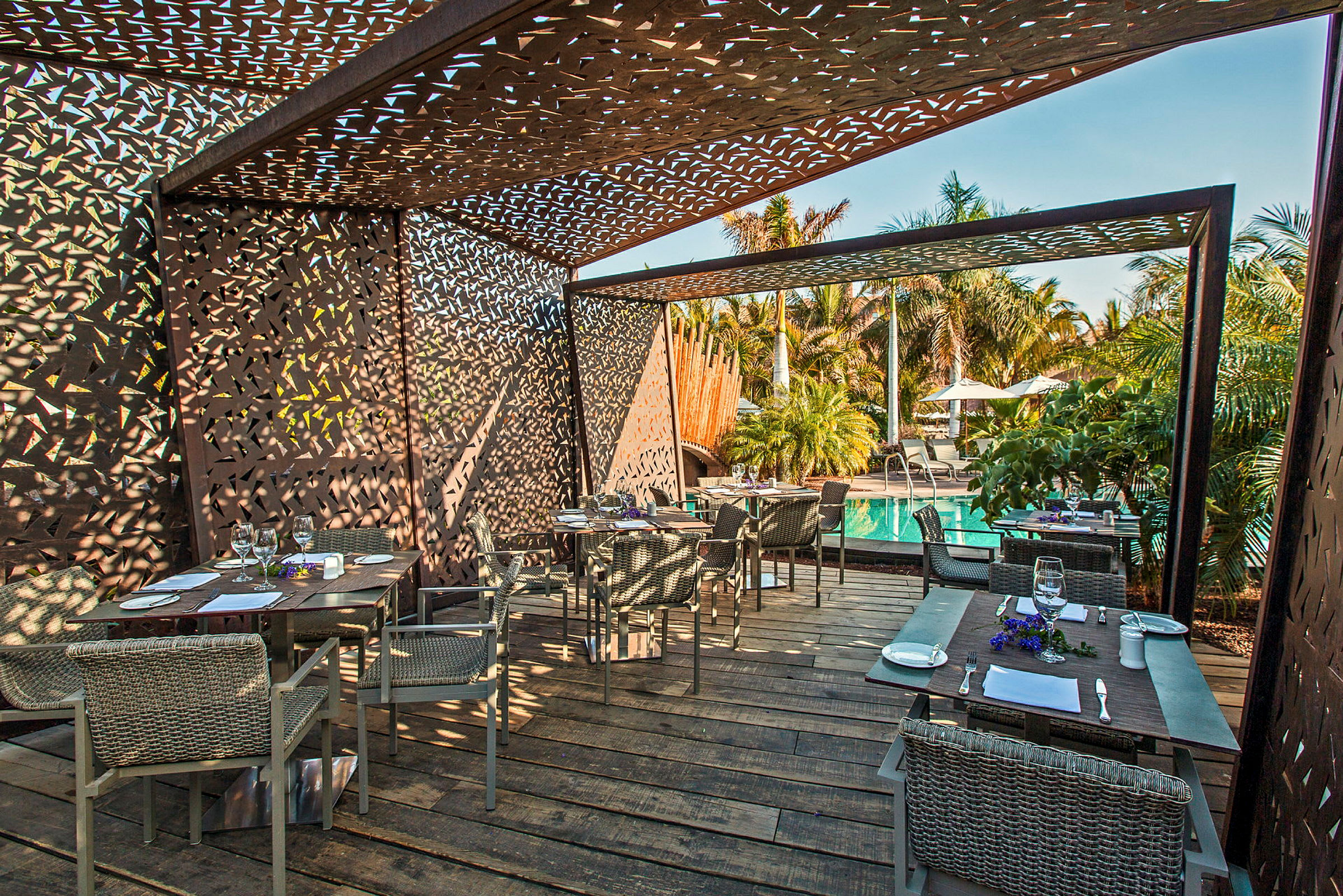 En udendørs restaurant med borde og stole under en skyggefuld struktur, ved siden af en pool og omgivet af palmer. (AI-genereret tekst)