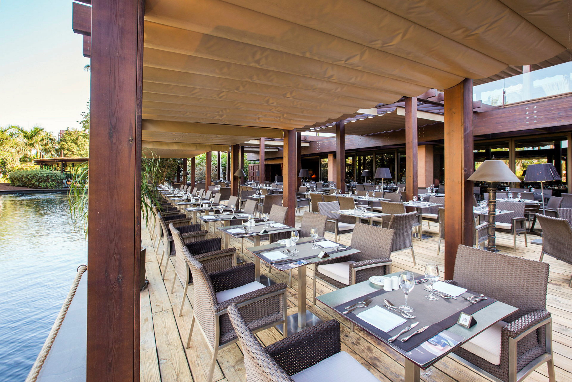 Billede af en terrasse med dækkede borde og stole under et beskyttende tag, placeret ved en vandoverflade. (AI-genereret tekst)