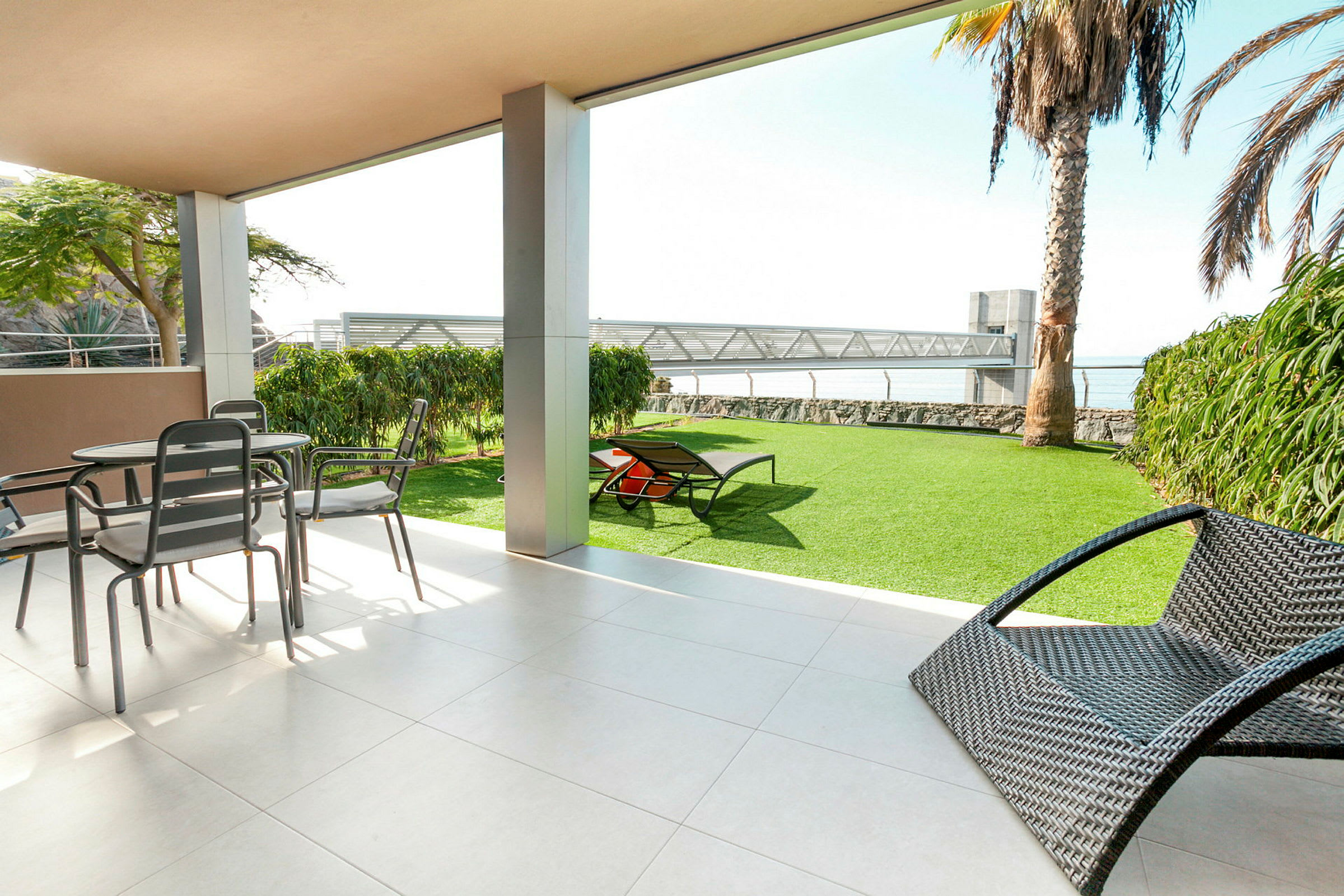Udendørs terrasse med bord og stole, solsenge på grøn græsplæne, udsigt mod havet. (AI-genereret tekst)