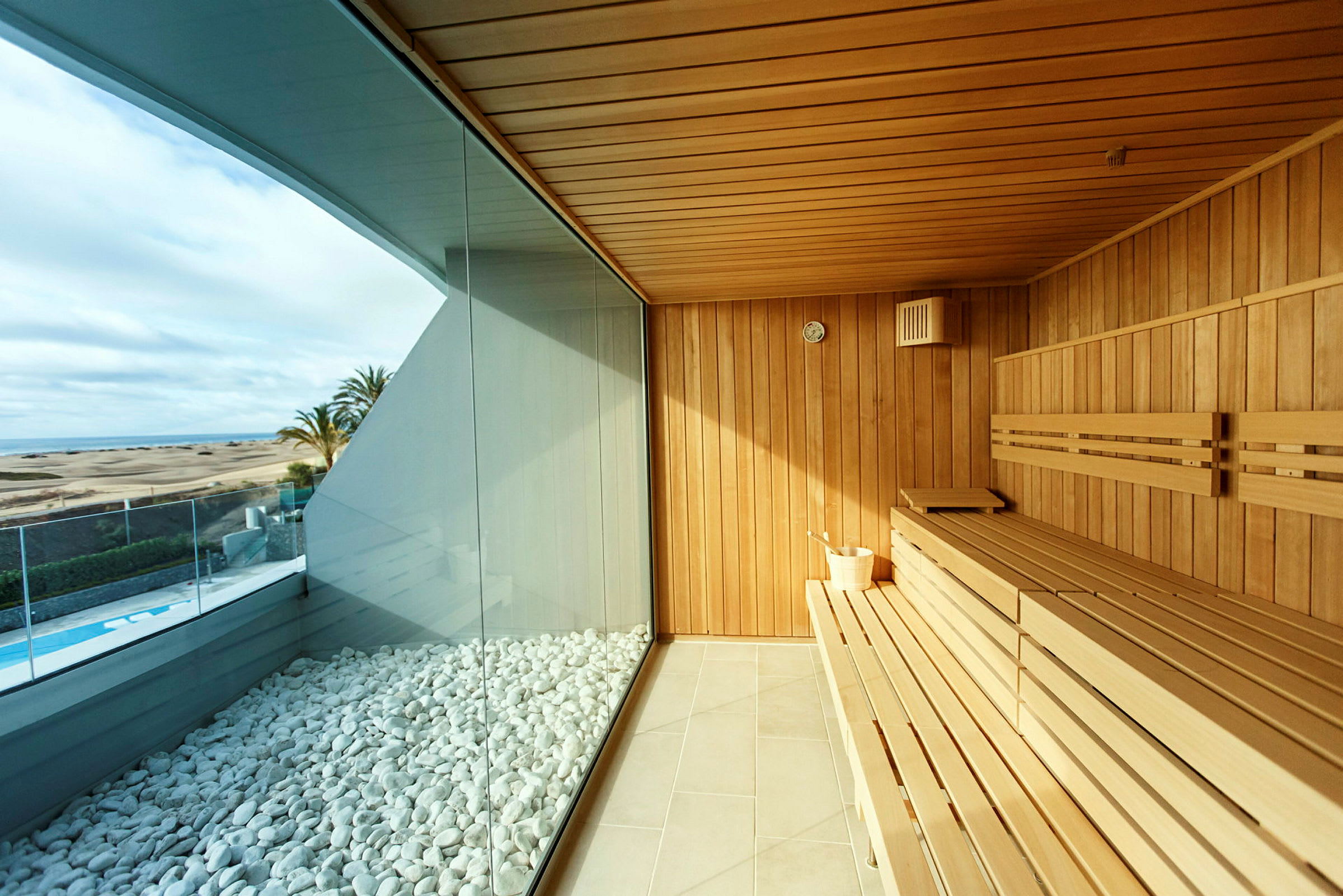 Indefra en sauna med træpaneler og store glasvinduer med udsigt til et poolområde og sandklitter. (AI-genereret tekst)