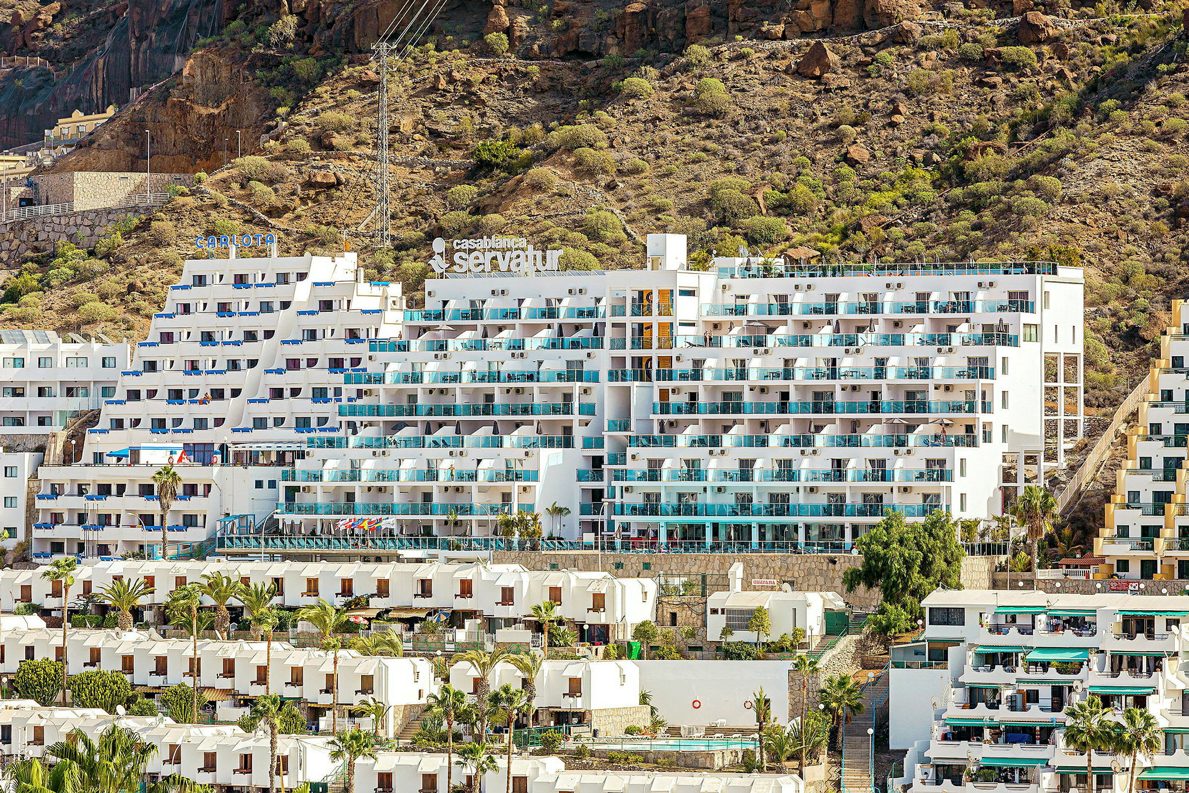 Billede af et hotel med hvid facade og altaner, beliggende ved en klippefyldt skråning. (AI-genereret tekst)