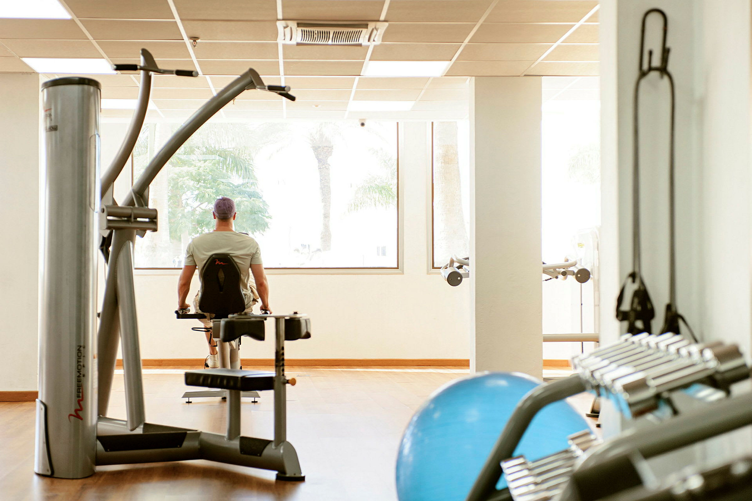 Et fitnessområde med træningsudstyr og en person, der bruger en maskine foran vinduer. (AI-genereret tekst)