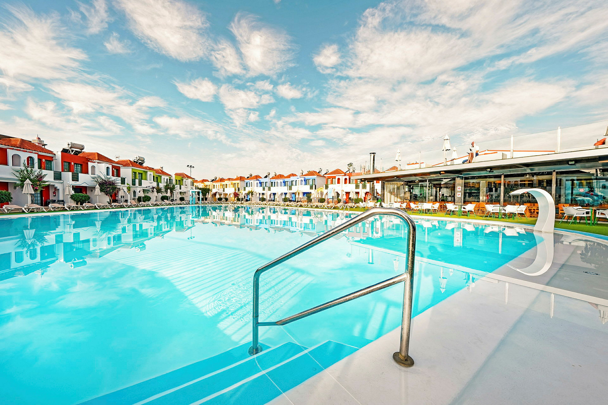 Udendørs pool omgivet af farverige bygninger og parasoller i roligt vejr. (AI-genereret tekst)