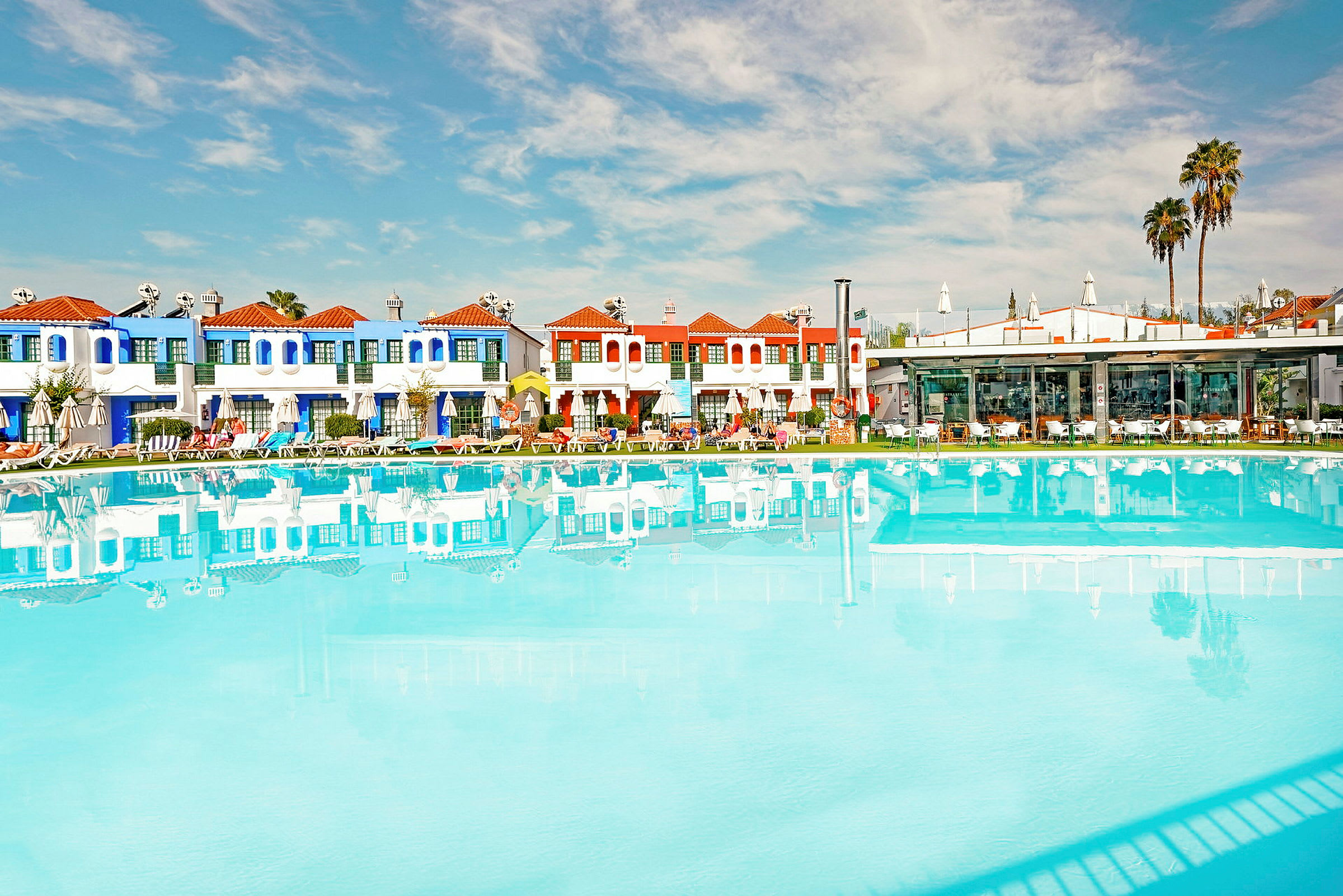 En swimmingpool foran en række farverige bygninger og palmer under en klar blå himmel. (AI-genereret tekst)