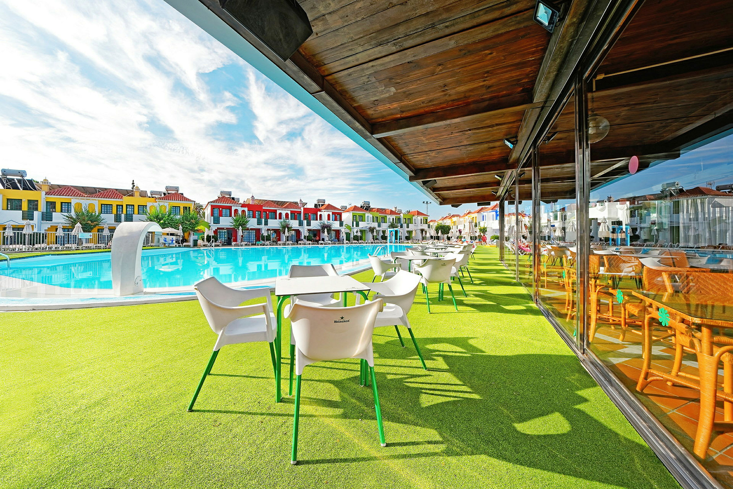 Udendørs serveringsområde med hvide stole og borde ved en pool, med farverige bygninger i baggrunden. (AI-genereret tekst)