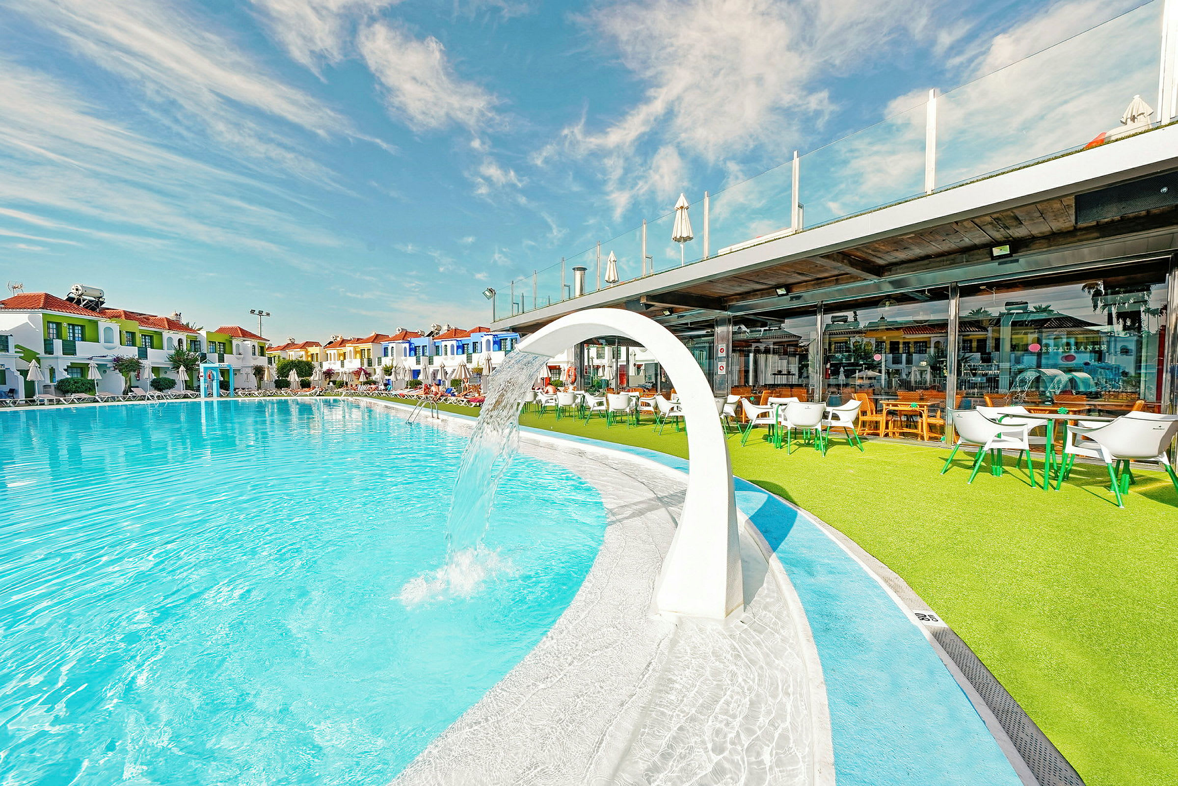 Stor swimmingpool med hvid vandbue, omgivet af farverige bygninger og et græsareal med udendørsmøbler. (AI-genereret tekst)