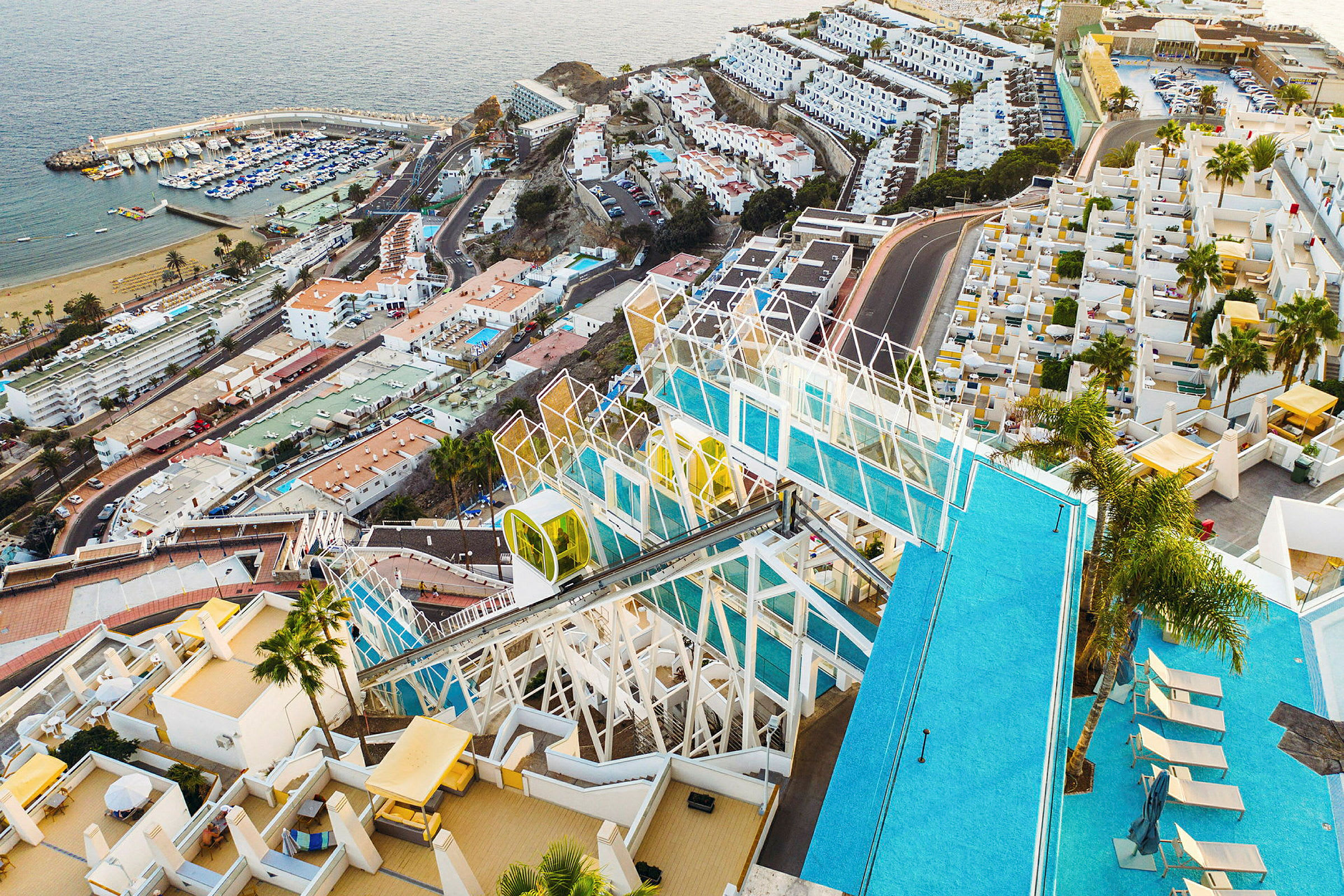 Udsigt over en pool og hotelområde med hav og småbådehavn i baggrunden. (AI-genereret tekst)
