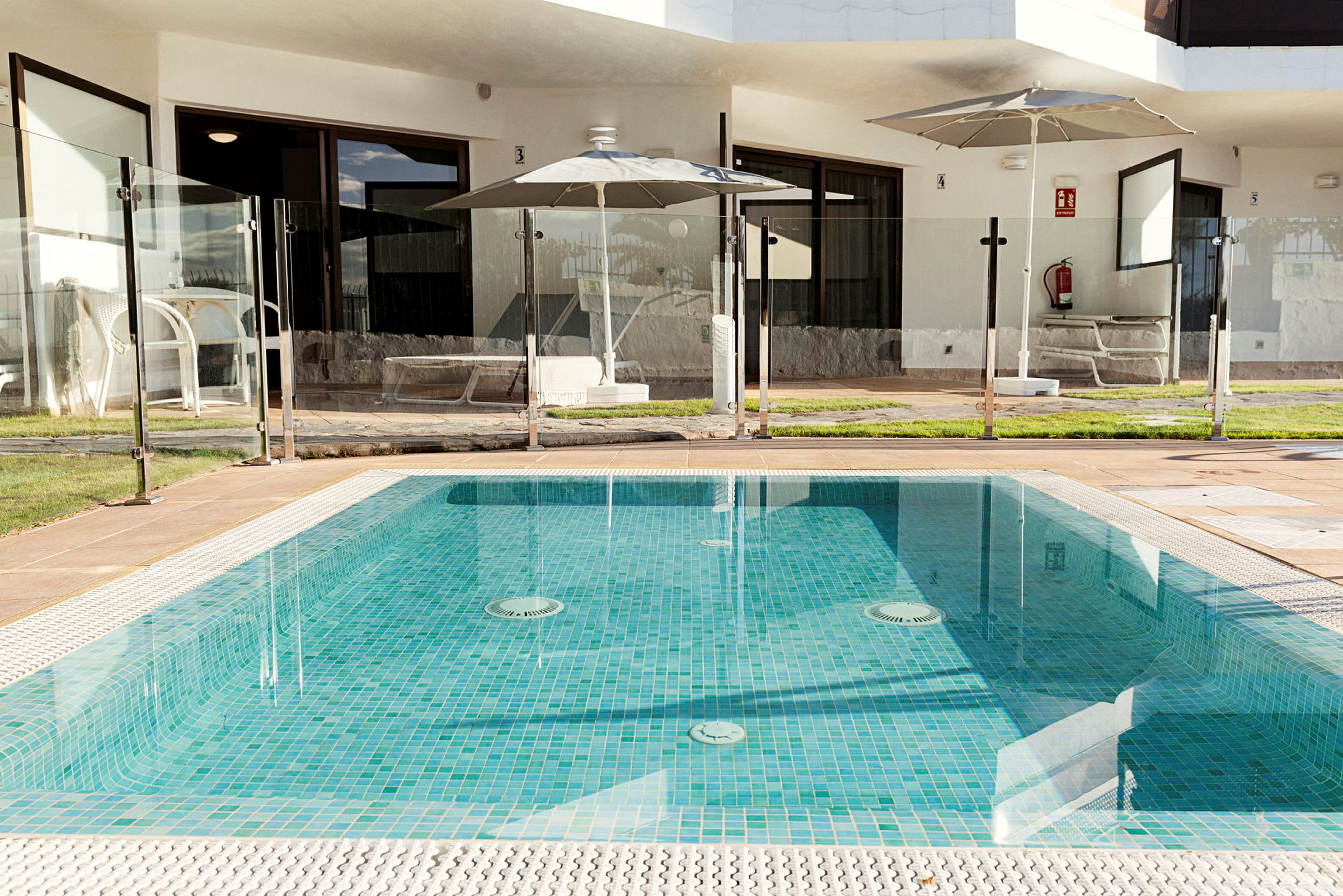 Udendørs pool med glasafskærmning og omgiverende græsplæne foran en hotelbygning. (AI-genereret tekst)