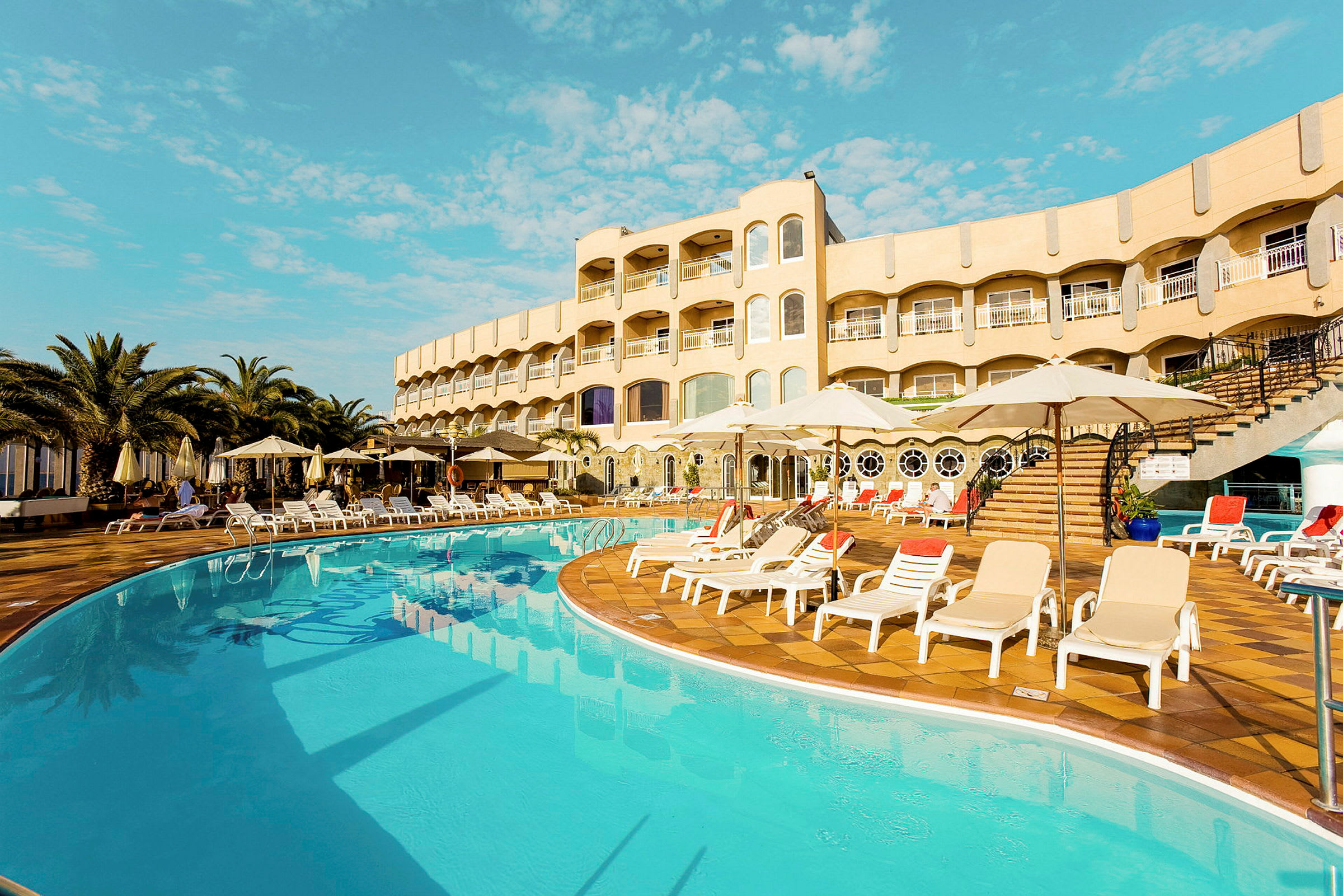 Udendørs pool med liggestole og parasoller foran en hotelbygning. (AI-genereret tekst)