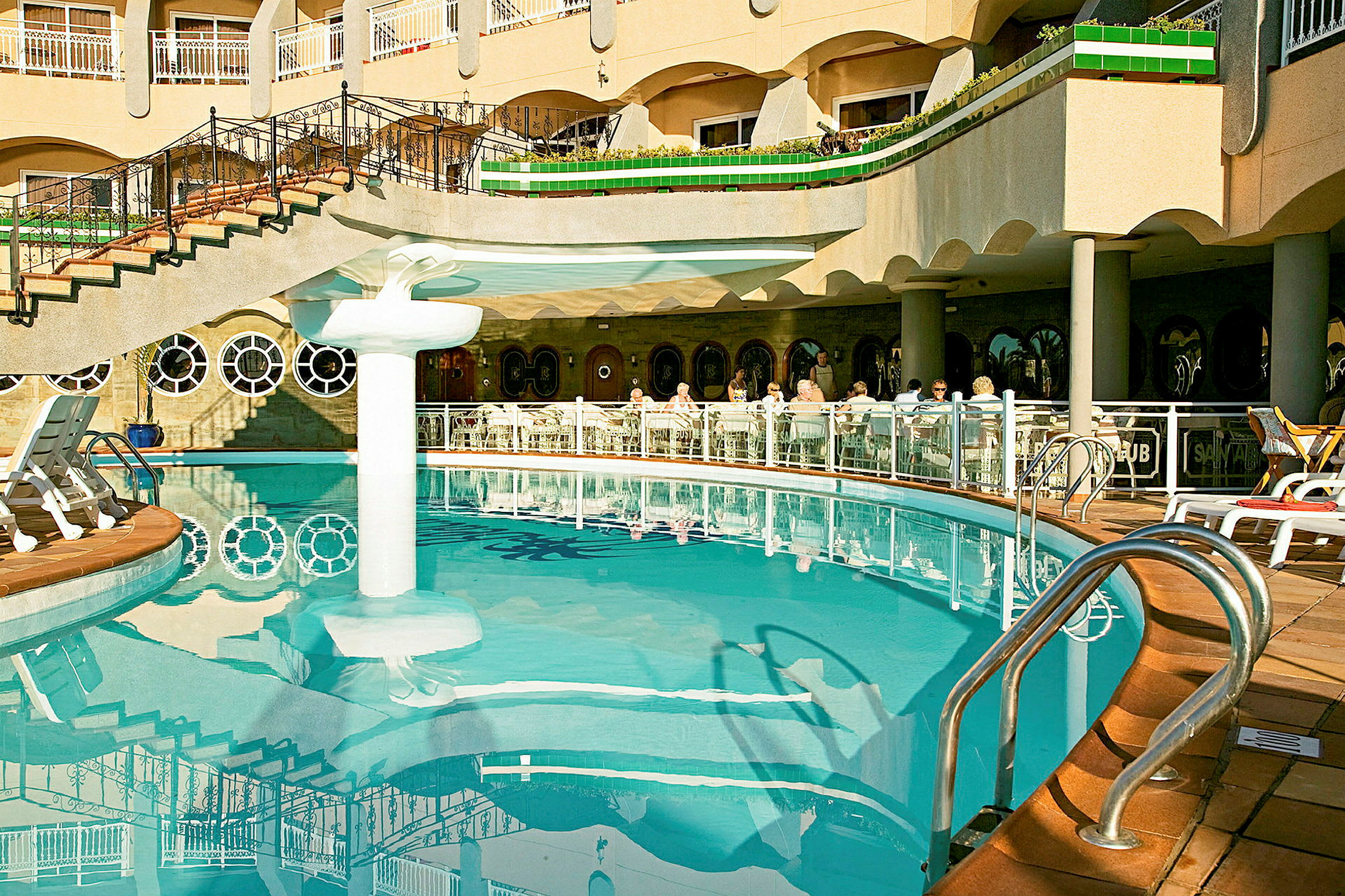 En udendørs pool ved et hotel med liggestole og gæster, der sidder ved borde. (AI-genereret tekst)