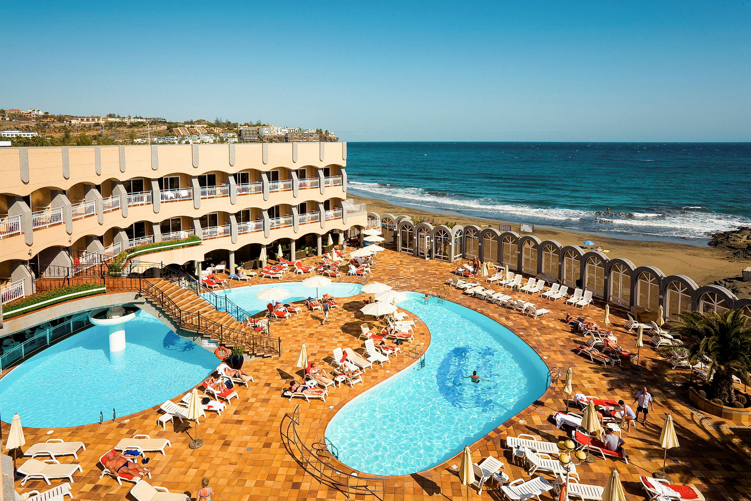 Hotelområde med pool og liggestole tæt på havet. (AI-genereret tekst)