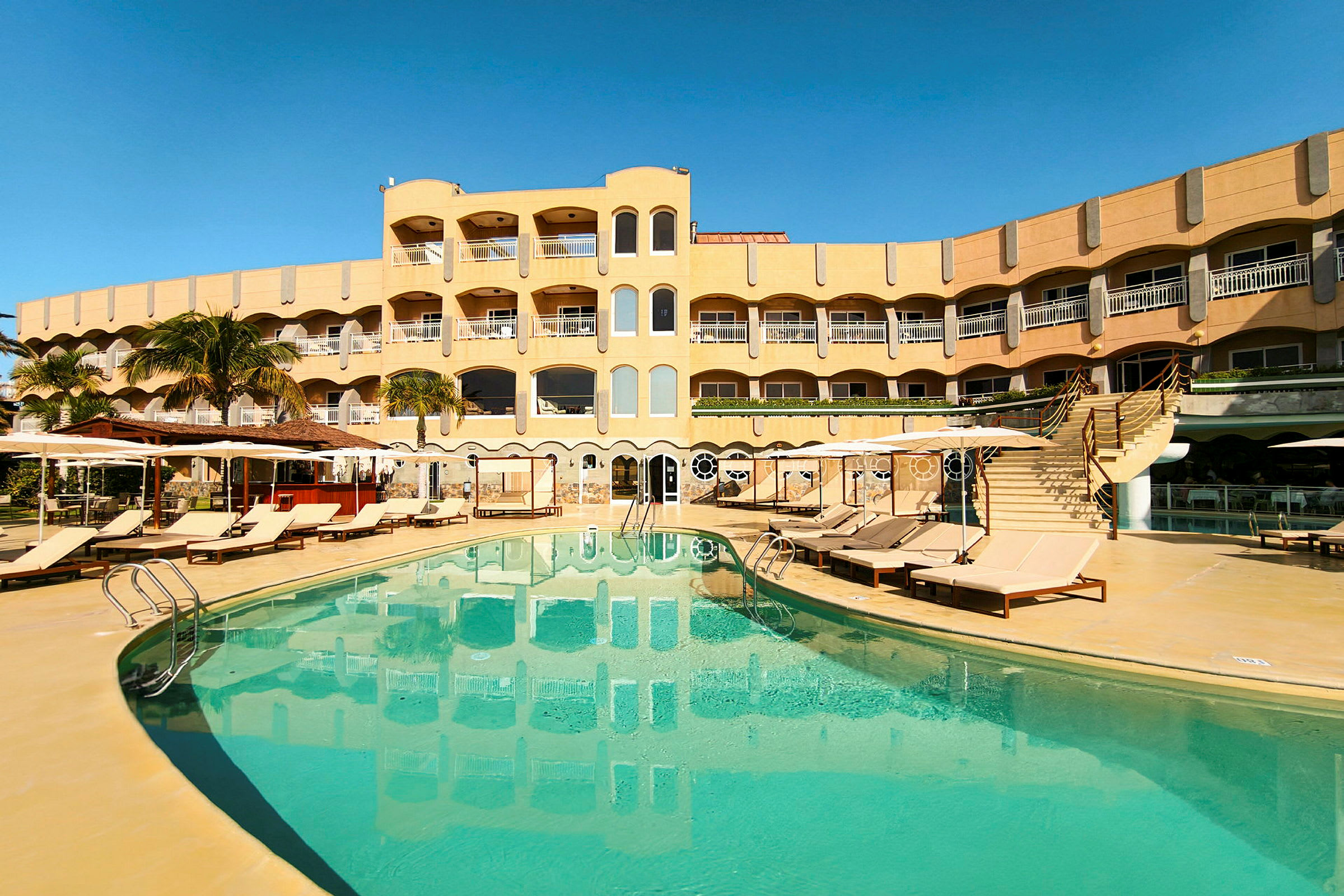 Et hotel med gul facade, altaner og en swimmingpool omgivet af liggestole og parasoller. (AI-genereret tekst)