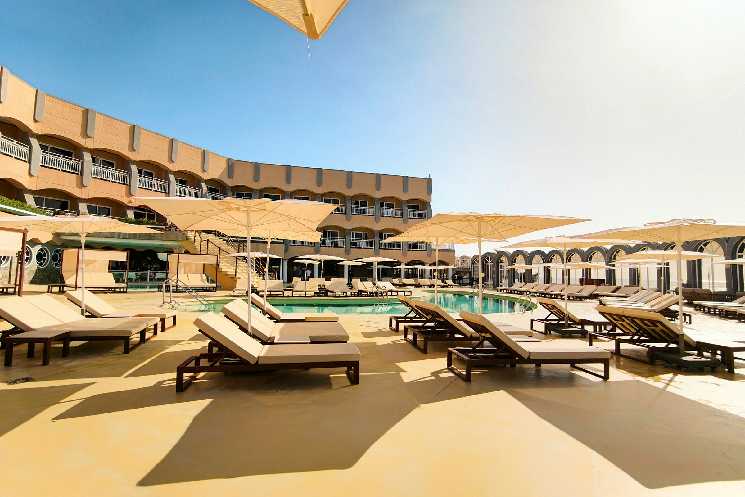 En udendørs pool med mange liggestole og parasoller ved et hotel. (AI-genereret tekst)
