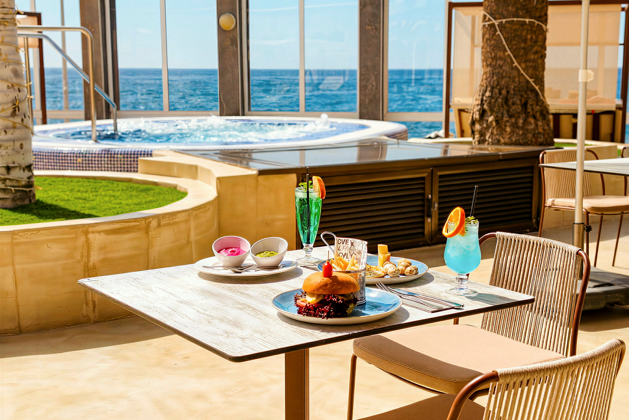 Et bord dækket med mad og drikke på en terrasse med havudsigt. I baggrunden ses en pool. (AI-genereret tekst)
