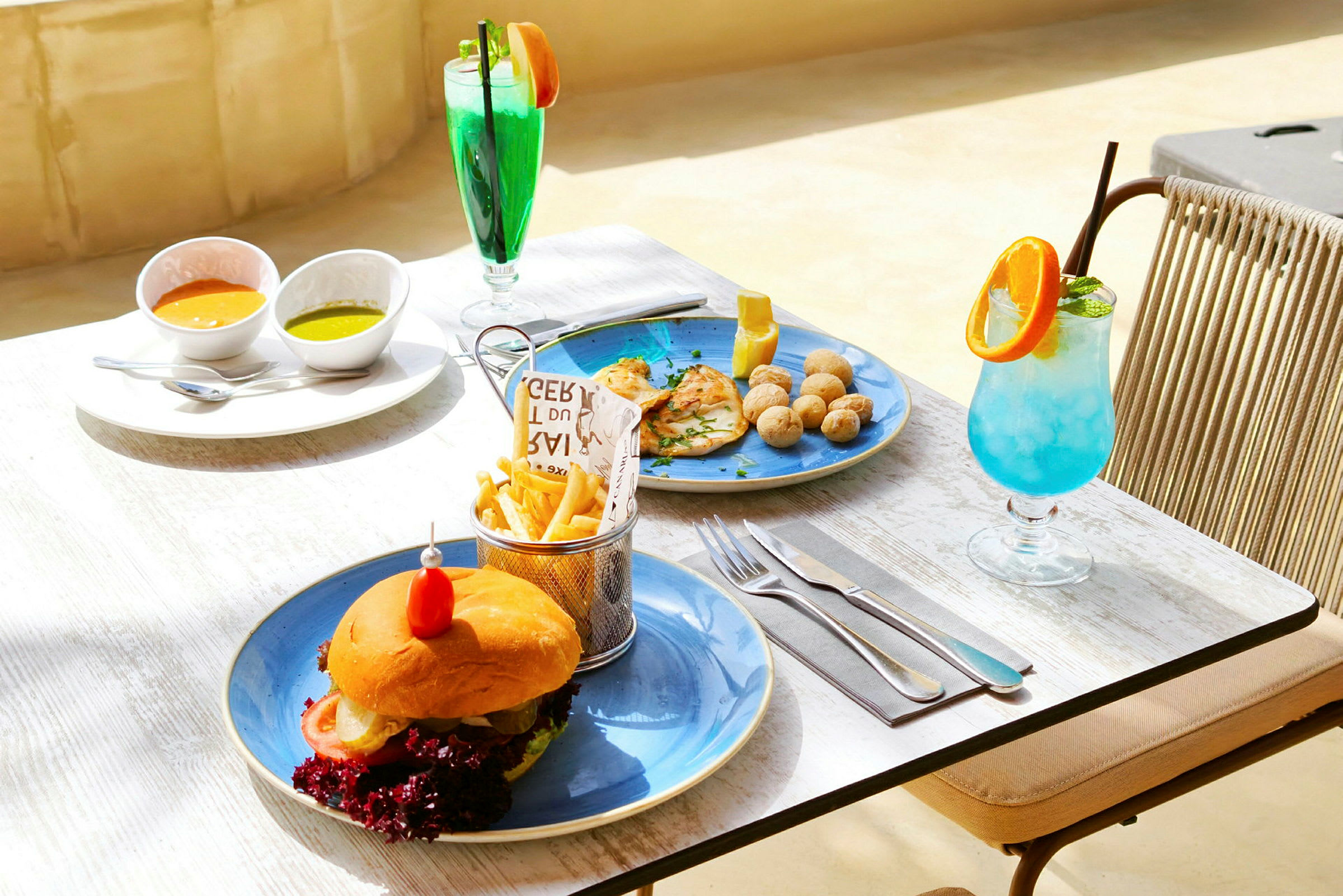 Et bord udendørs med tallerkener der serverer en burger med pommes frites, to skåle med dip-saucer, en ret med kylling og kartofler samt to farverige drinks. (AI-genereret tekst)