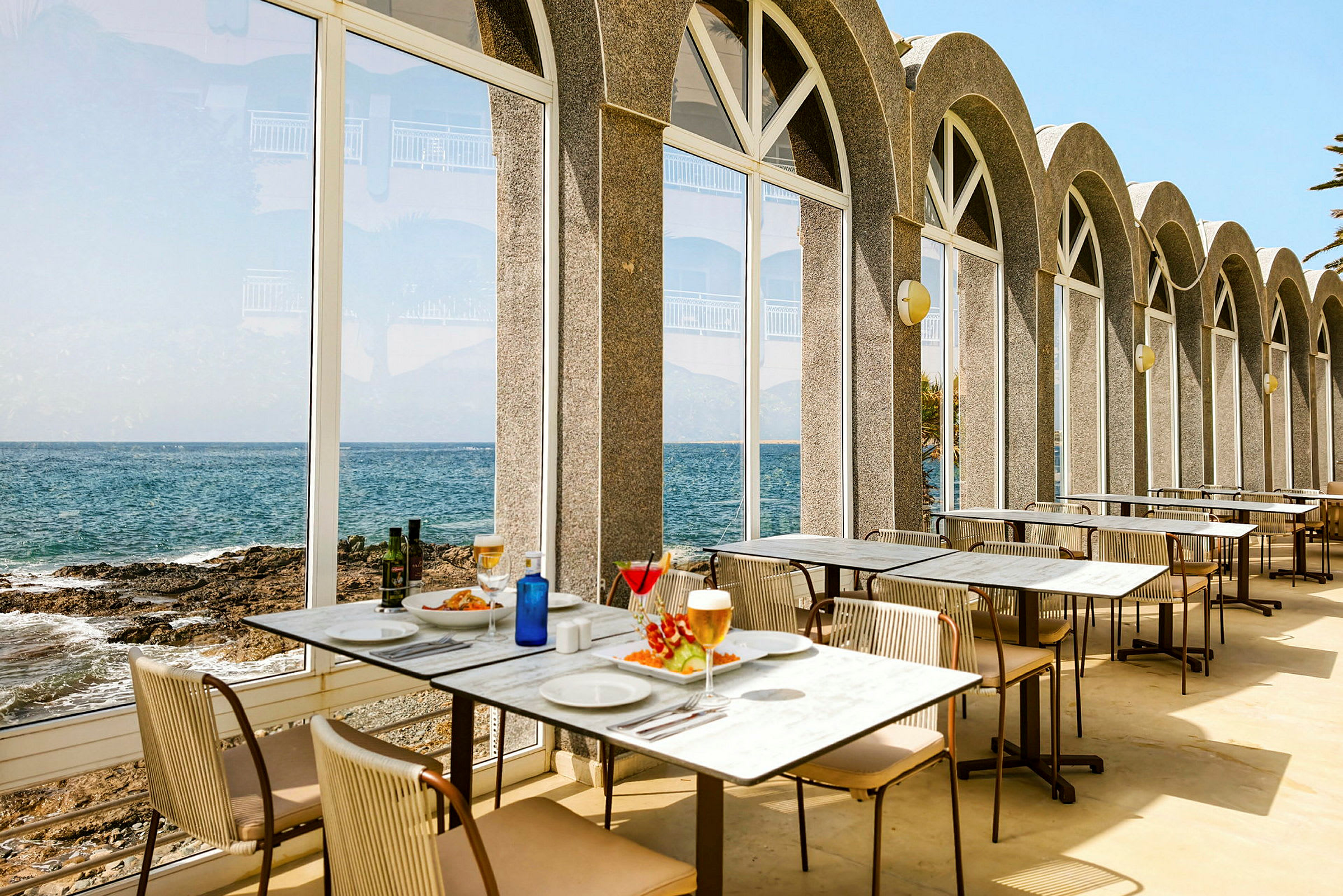 Restauranter med borde opstillet ved store glasvinduer, der vender ud mod havet. (AI-genereret tekst)