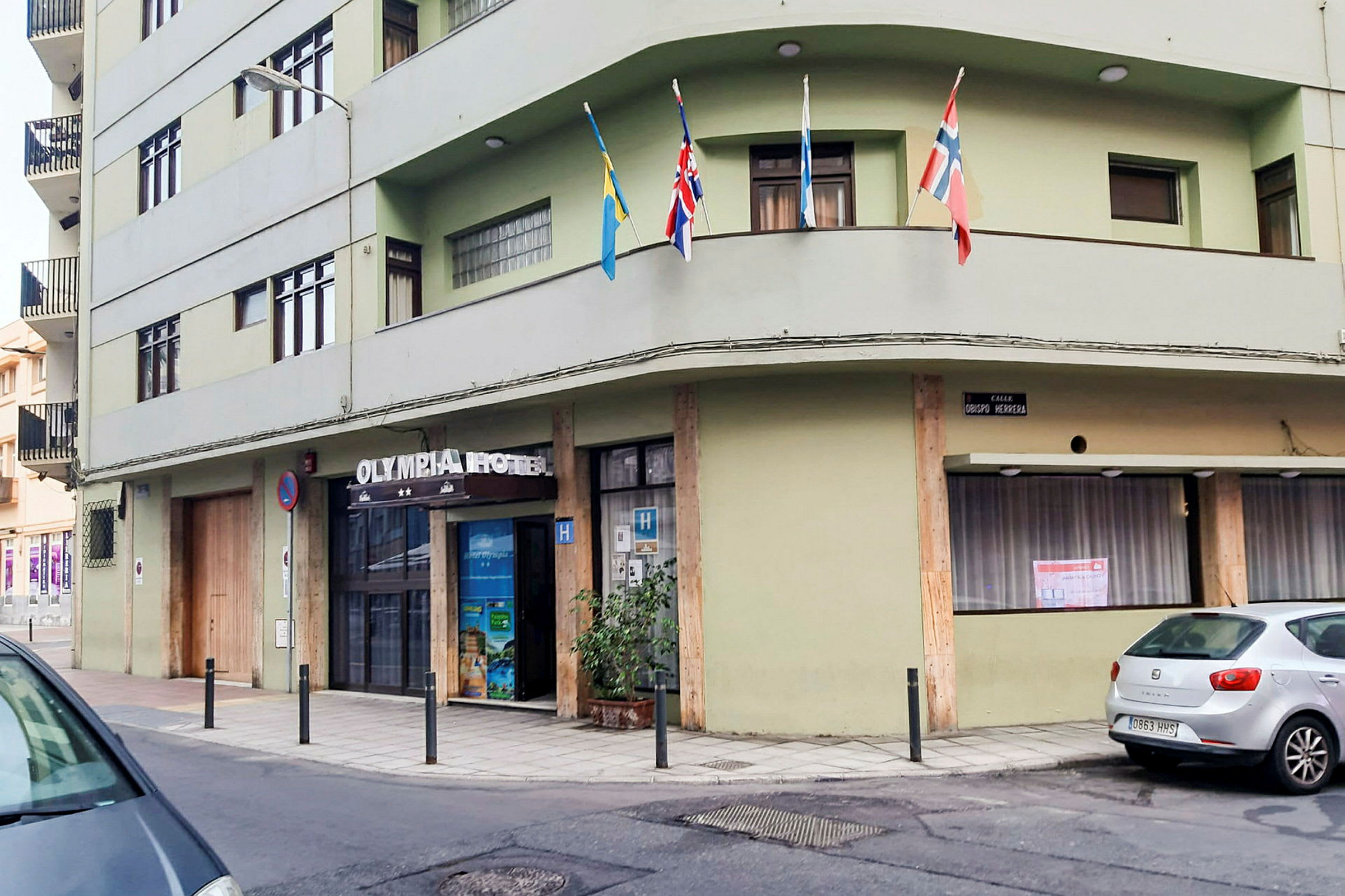 En grøn bygning med teksten 'Olympia Hotel' over indgangen. Flere flag er opsat på en altan. (AI-genereret tekst)