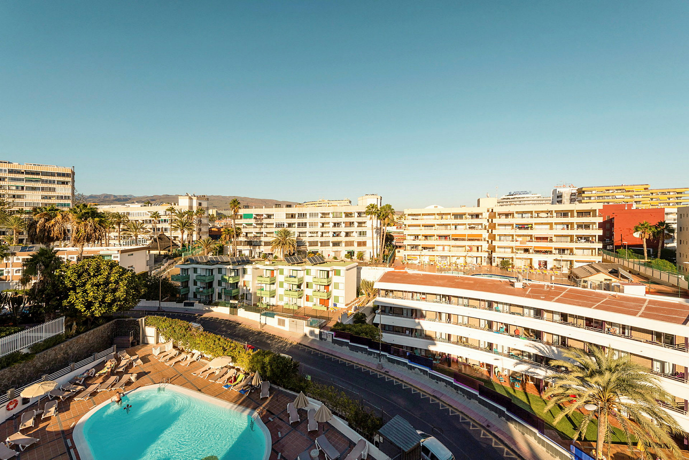 Udsigt over et hotelområde med pool, omgivet af bygninger under en klar himmel. (AI-genereret tekst)