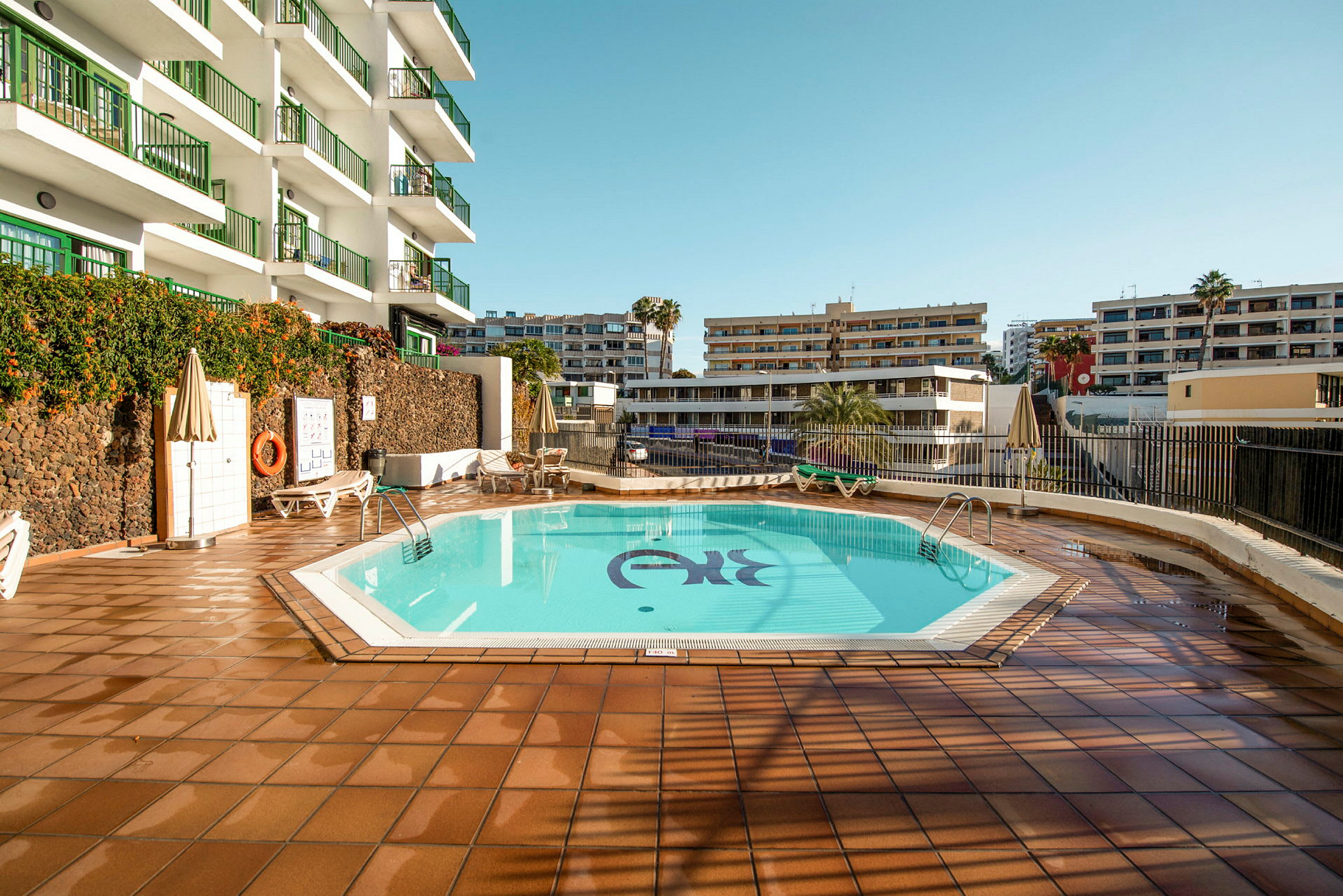 En hotelpool omgivet af liggestole og bygninger. (AI-genereret tekst)