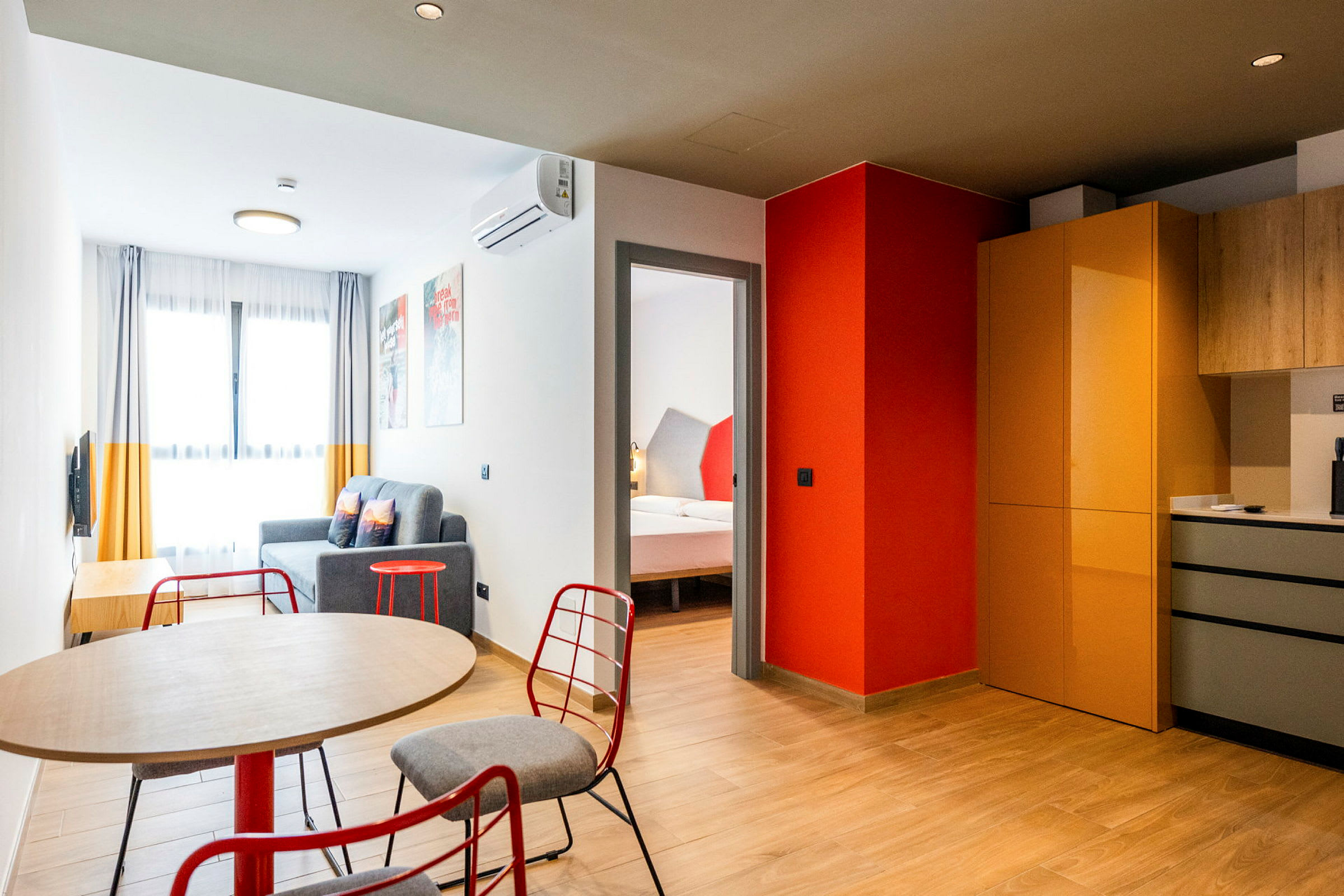 Et moderne indrettet hotelværelse med et bord og stole i forgrunden, en siddegruppe med sofa og et soveværelse synligt gennem en døråbning. Køkkenområdet har skabe og arbejdsområder. (AI-genereret tekst)