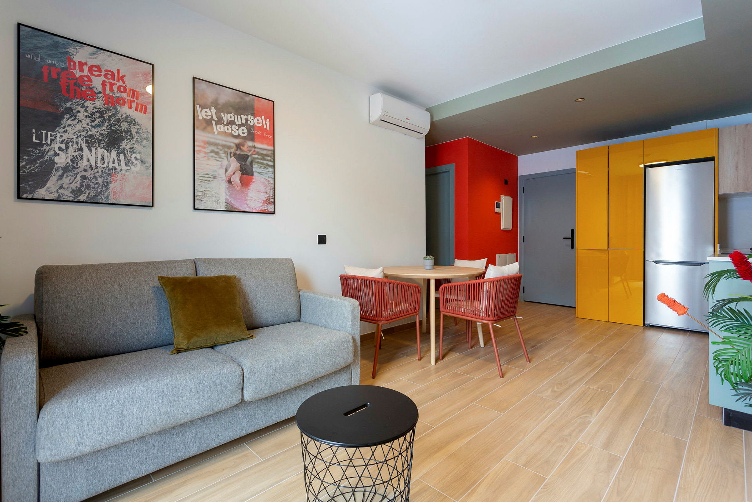 En moderne stue med en grå sofa og et lille sort bord. På væggen er der to farverige malerier. Til højre ses et køkken med gul køkkenopbevaring og et køleskab. (AI-genereret tekst)