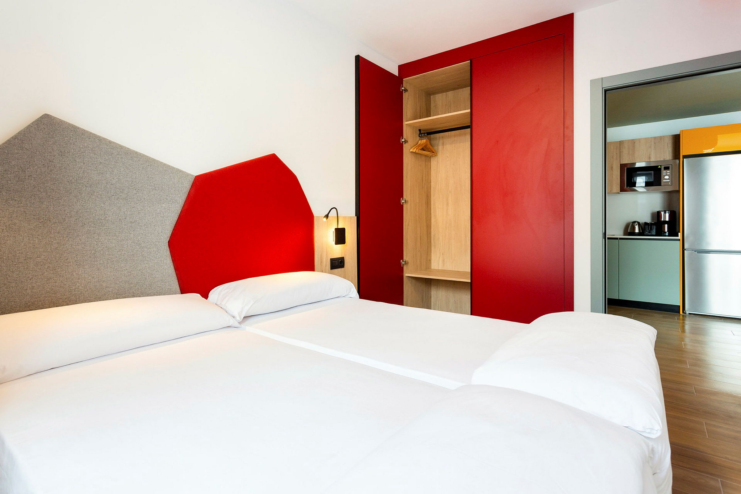 Billede af et moderne hotelværelse med en dobbeltseng, et skab med røde skydedøre og et åbent lille køkkenområde med køleskab og mikrobølgeovn. (AI-genereret tekst)