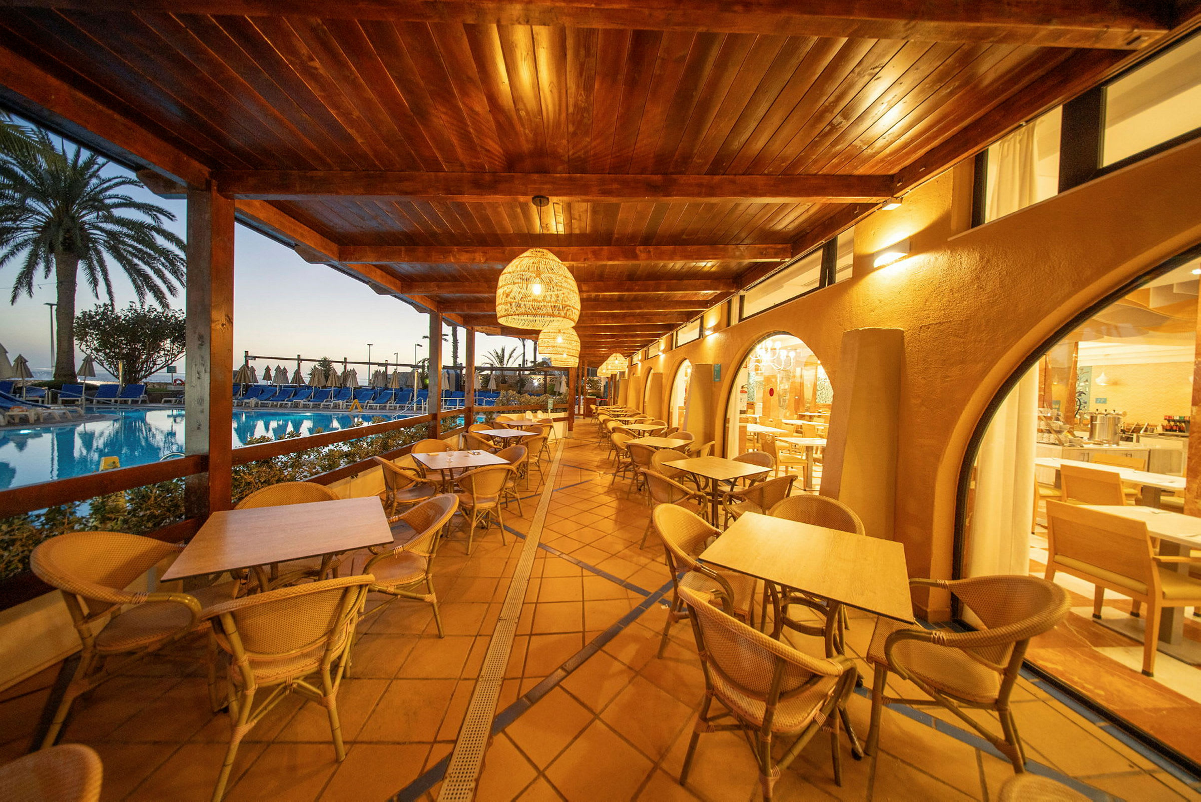 En udendørs terrasse med siddepladser og borde under et tag, ved siden af en pool og med udsigt over et område med liggestole og palmer. (AI-genereret tekst)