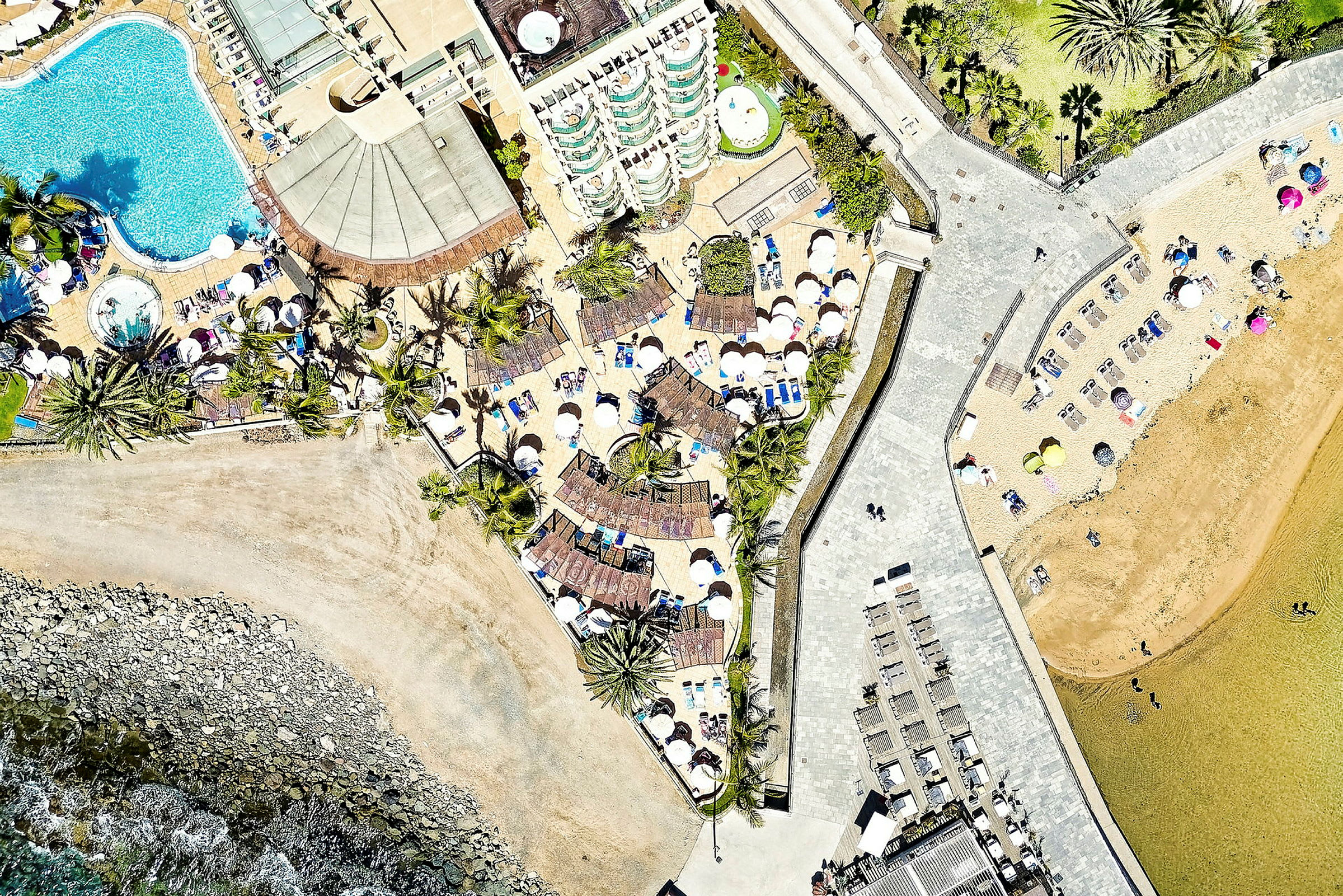Luftfoto af et feriested med swimmingpools, liggestole og parasoller tæt på stranden. (AI-genereret tekst)