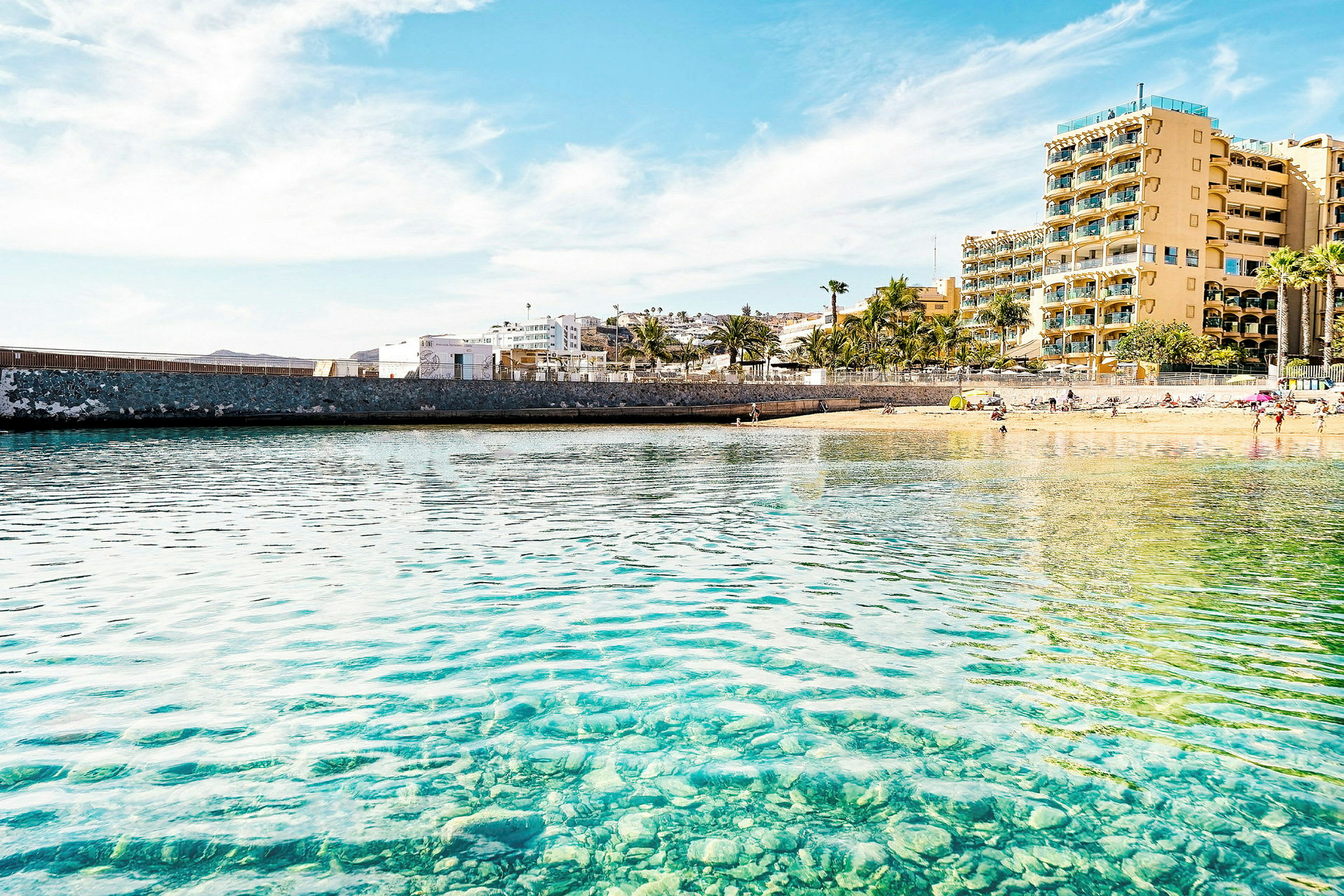 Strand med en stenmur og hotel i baggrunden. (AI-genereret tekst)