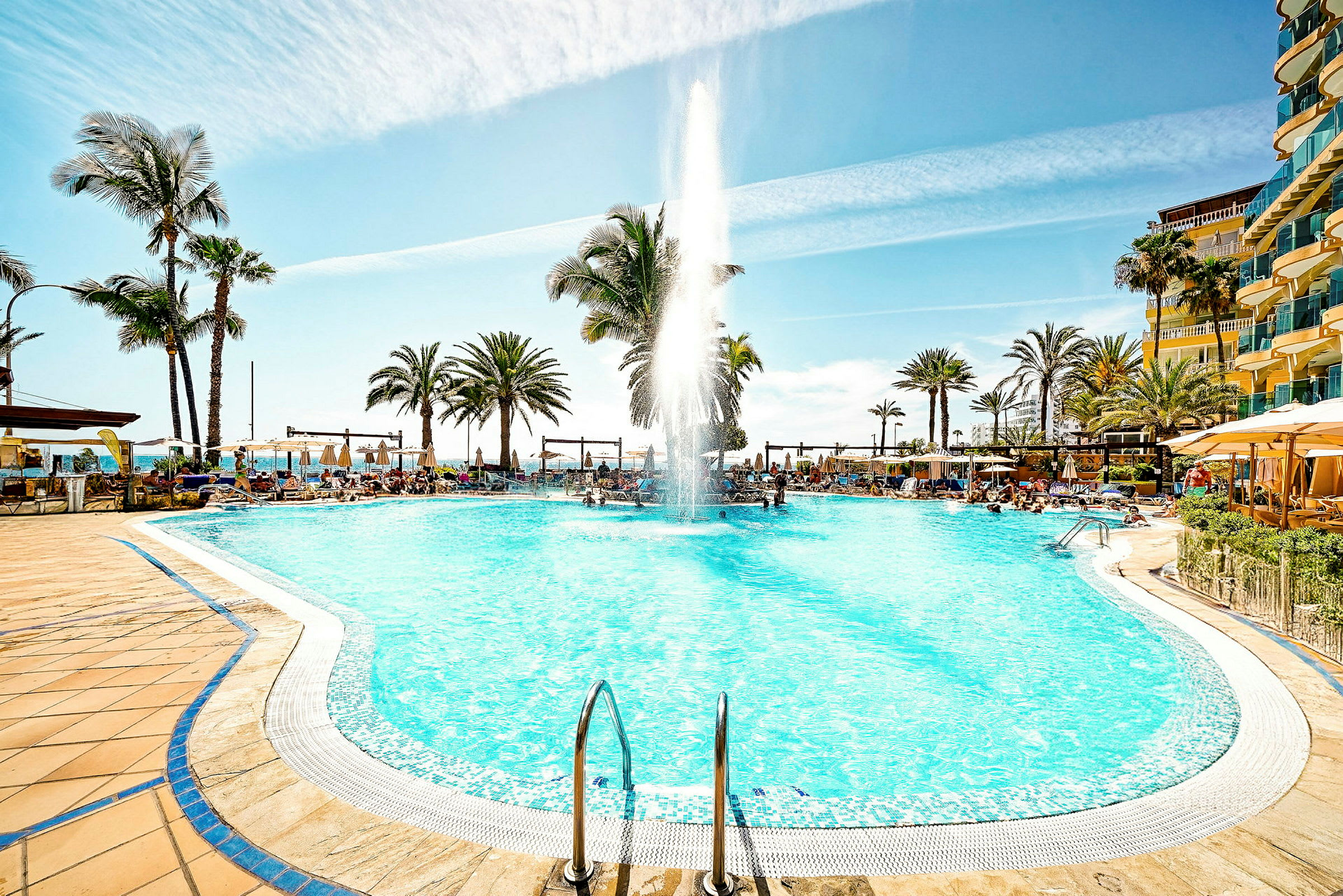 Poolområde med springvand omgivet af liggestole og palmer. (AI-genereret tekst)