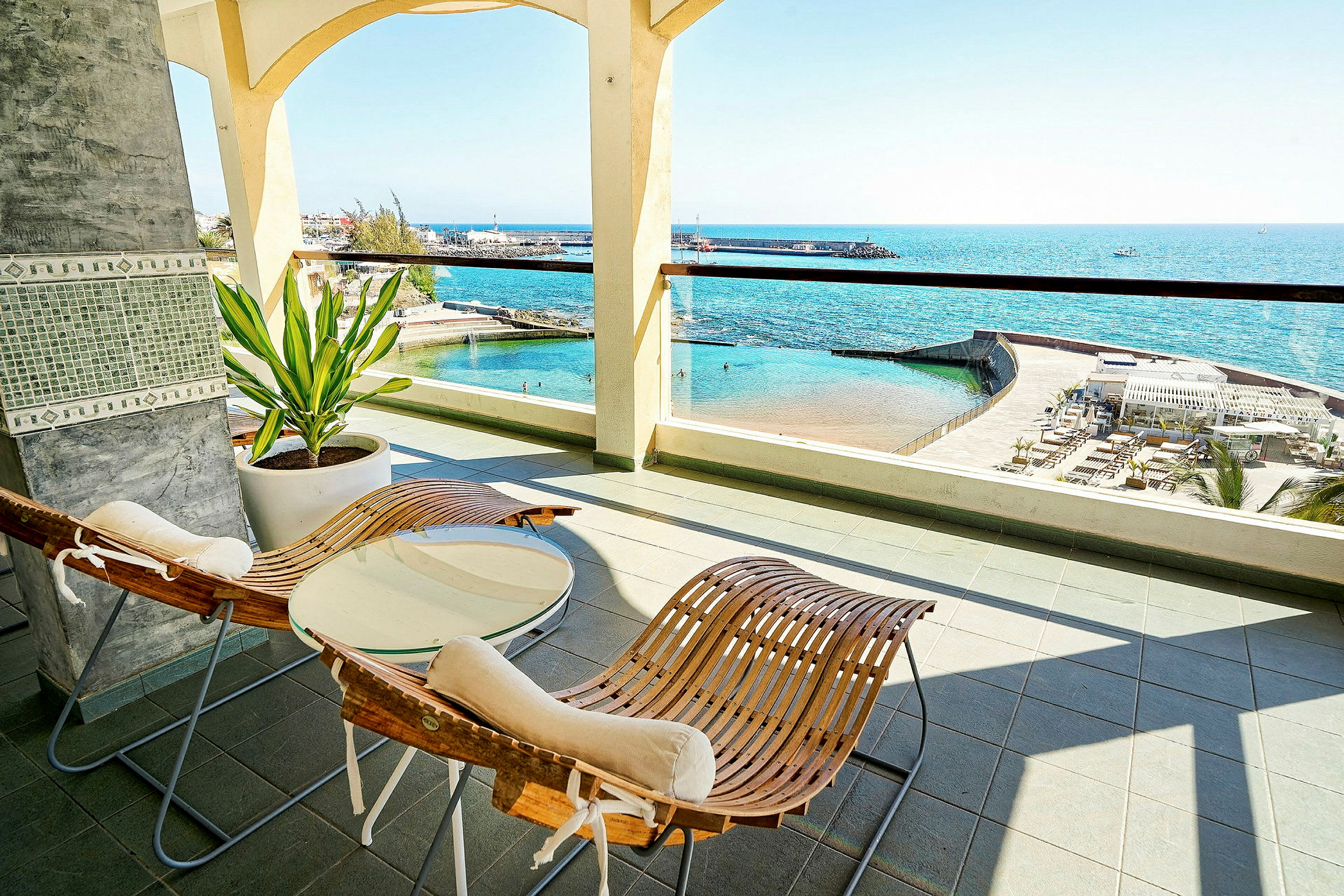 Terrasse med to træstole, et bord og udsigt over havet og en pool. (AI-genereret tekst)