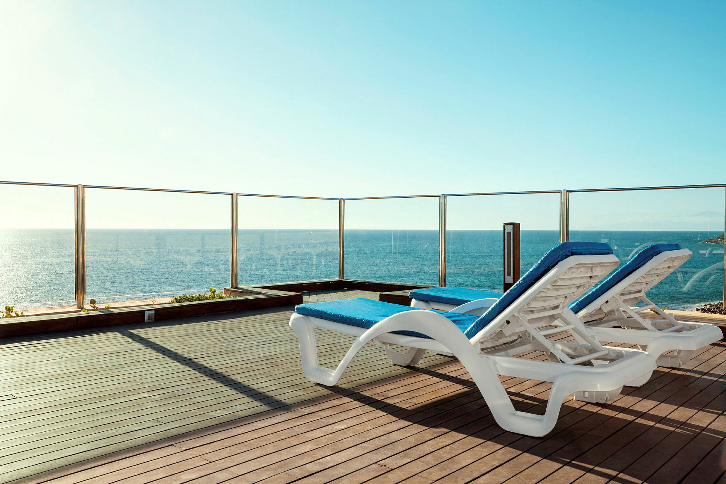 To liggestole med blå puder står på en terrasse med glasrækværk og udsigt over havet. (AI-genereret tekst)