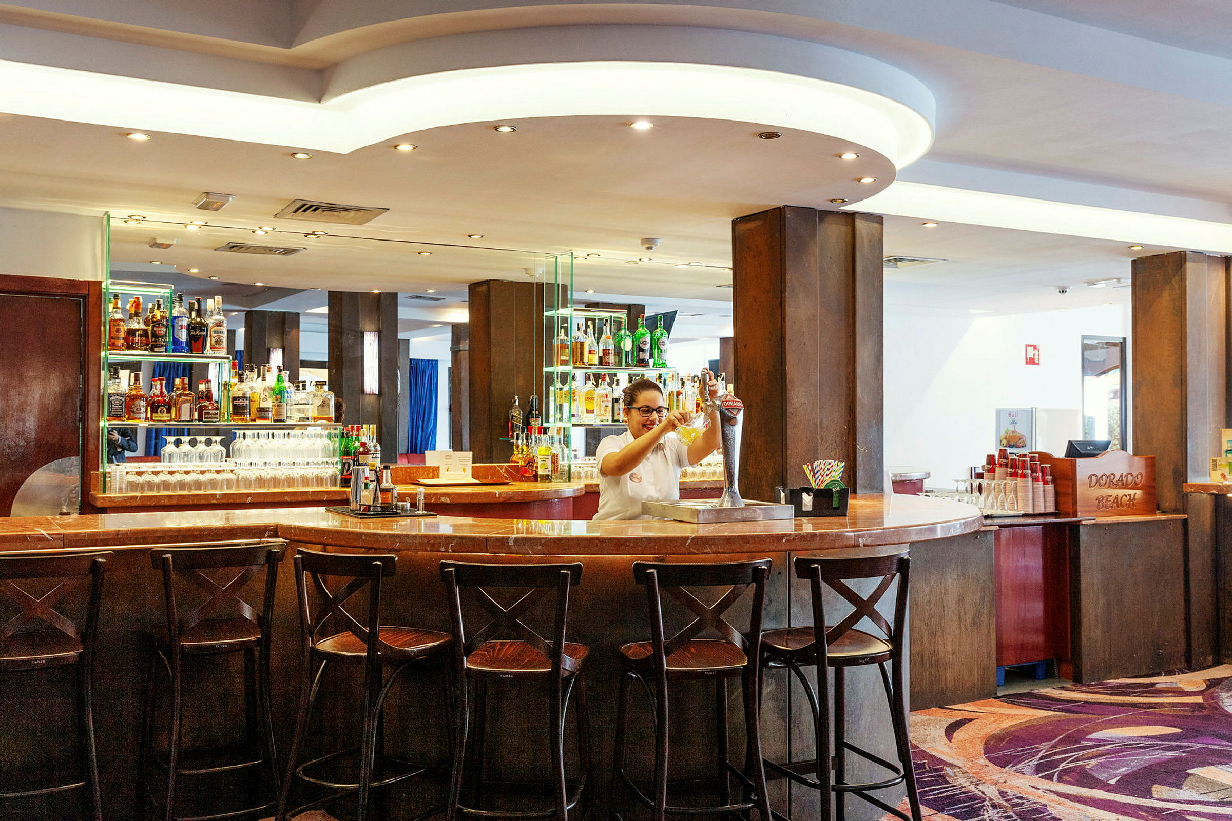 Billedet viser en velbelyst hotelbar med mørkt træinteriør og flere stole ved bardisken. (AI-genereret tekst)