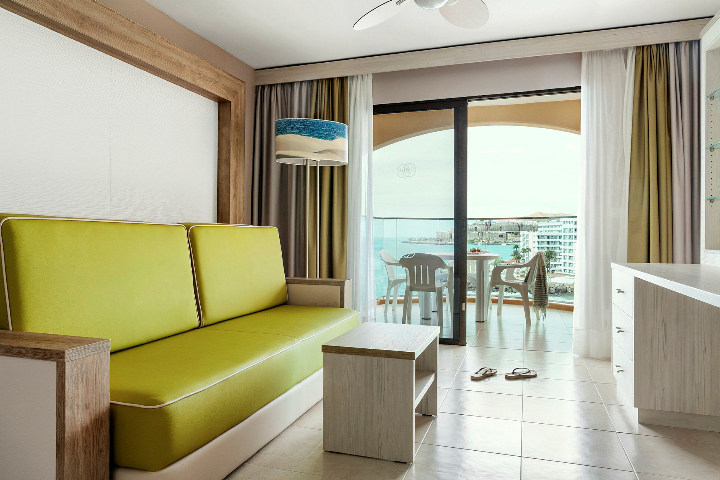 Interiør af et hotelværelse med en grøn sofa, et bord og vinduer der vender ud mod en balkon med udsigt over havet. (AI-genereret tekst)