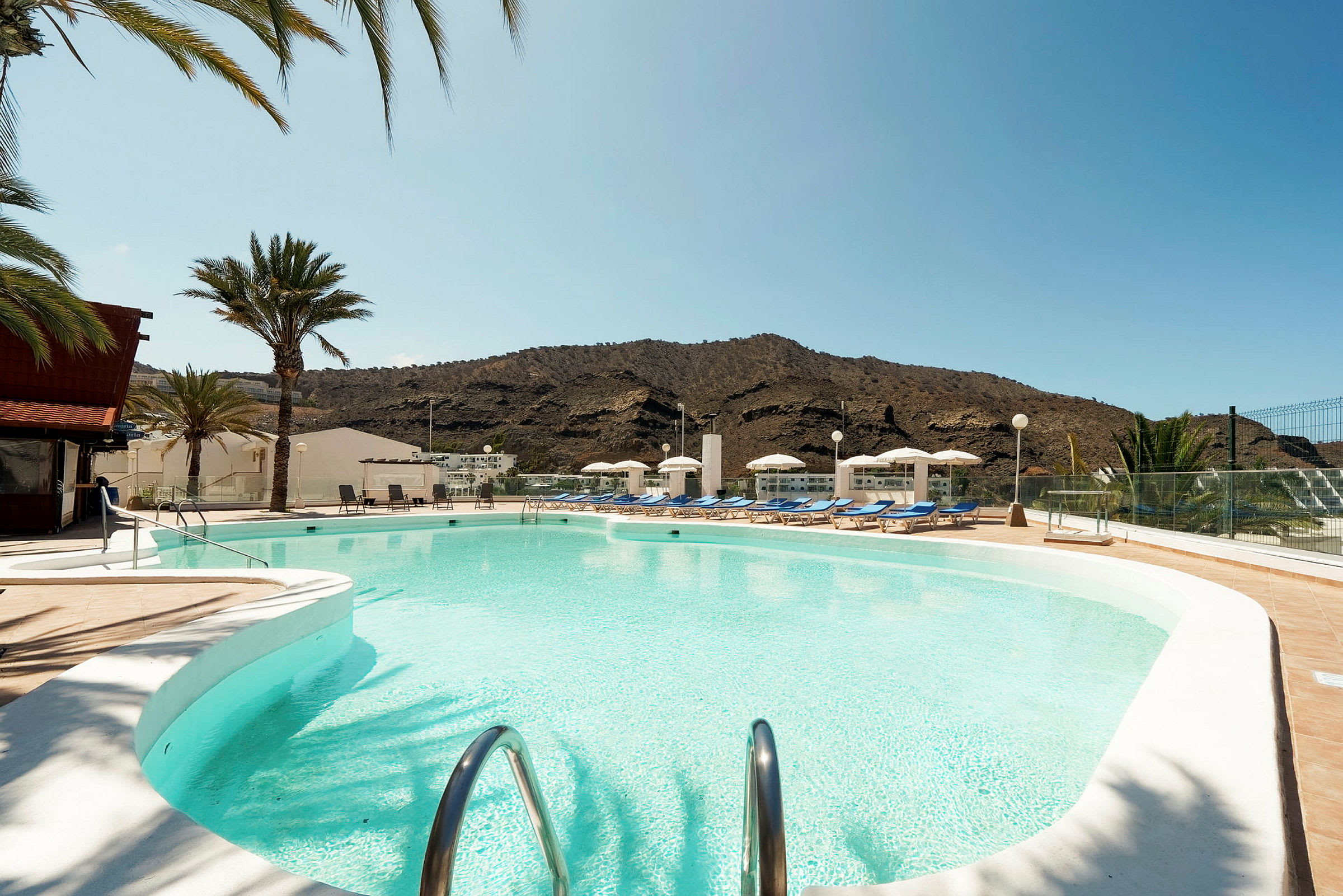 Udendørs swimmingpool med liggestole og parasoller, omgivet af palmer og bjerge. (AI-genereret tekst)
