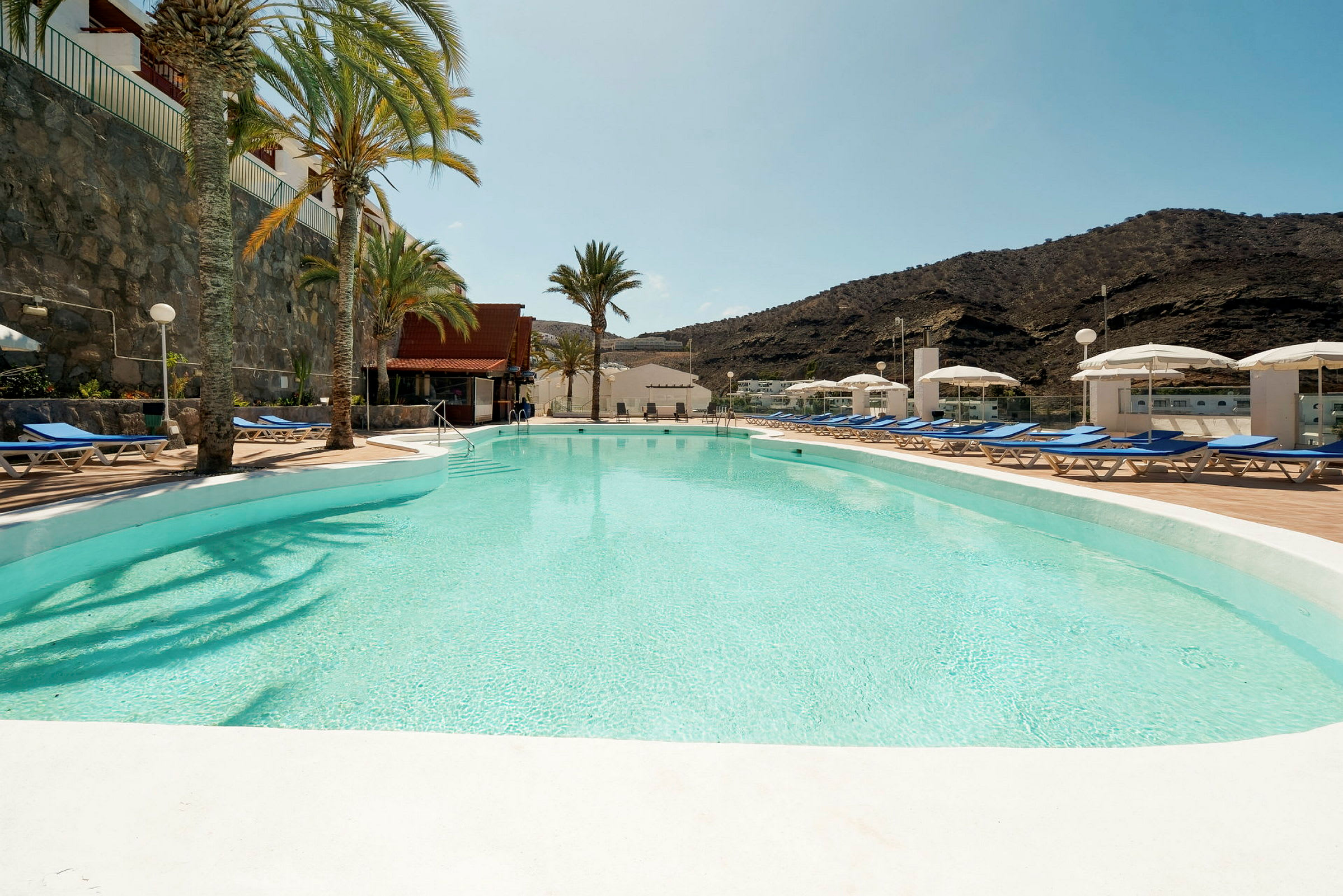 En udendørs pool med liggestole og parasoller ved siden af samt bjerge i baggrunden. (AI-genereret tekst)